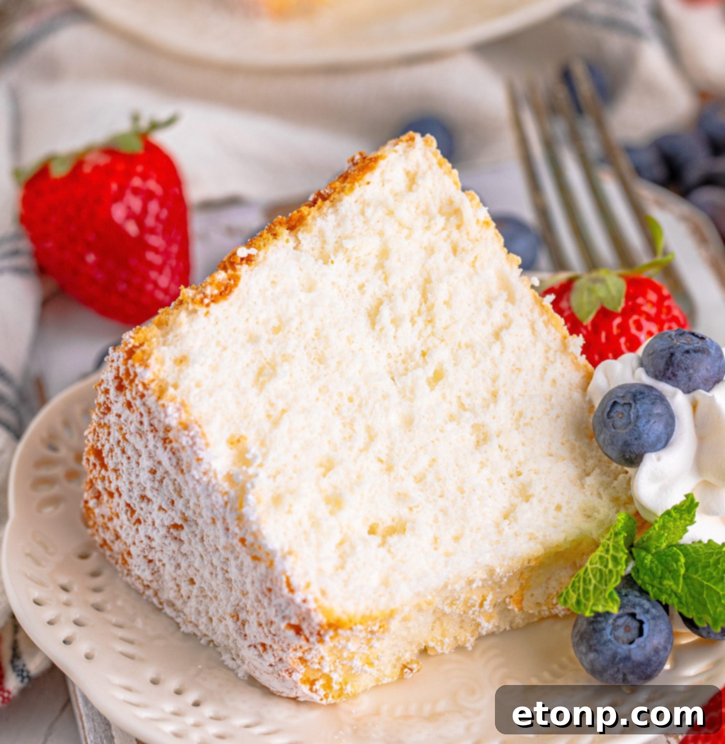 Close up of a slice of Angel Food Cake on a white plate with whipped cream, fresh berries, and mint