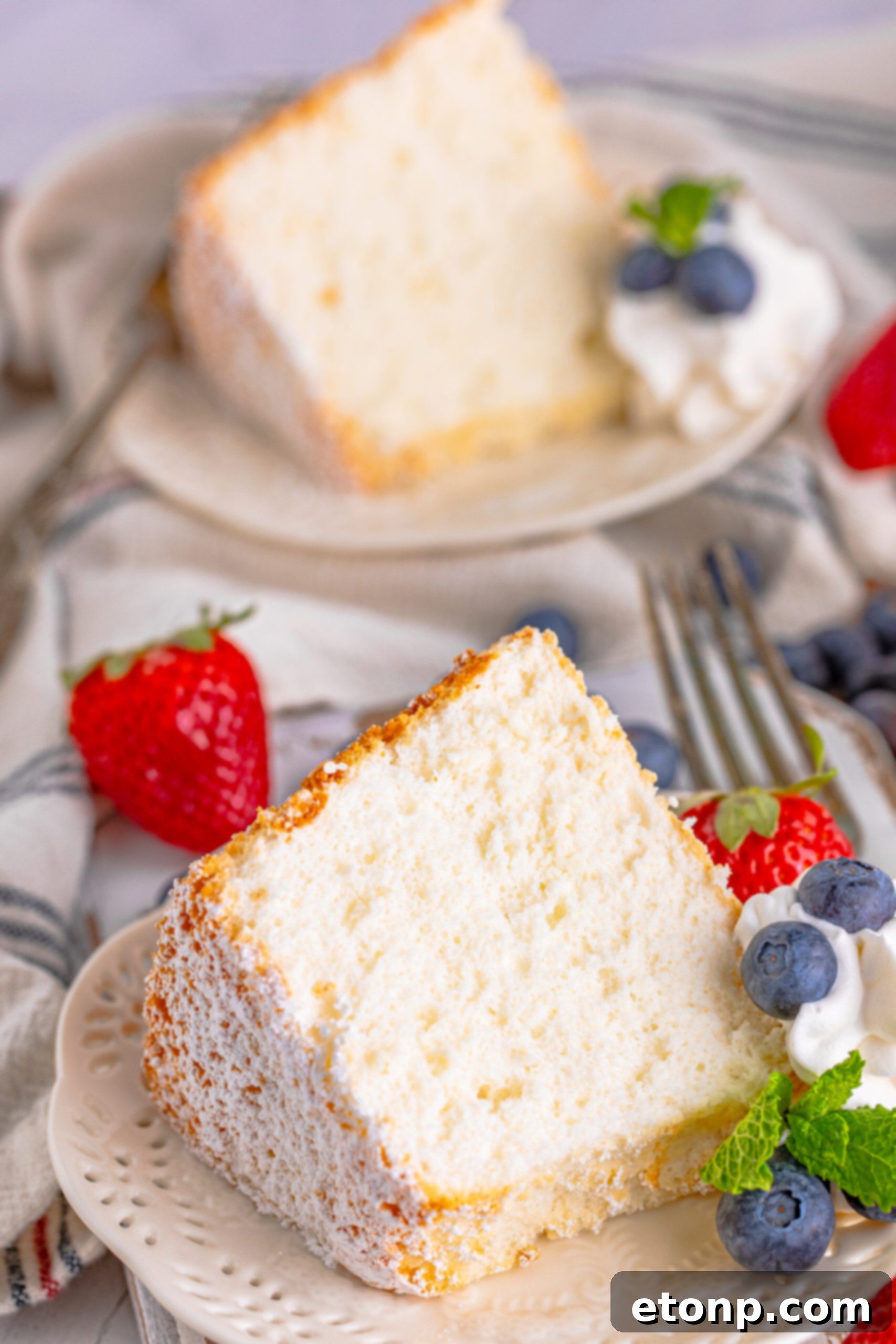 A piece of Angel Food Cake on a white plate with whipped cream, fresh berries, and mint. 