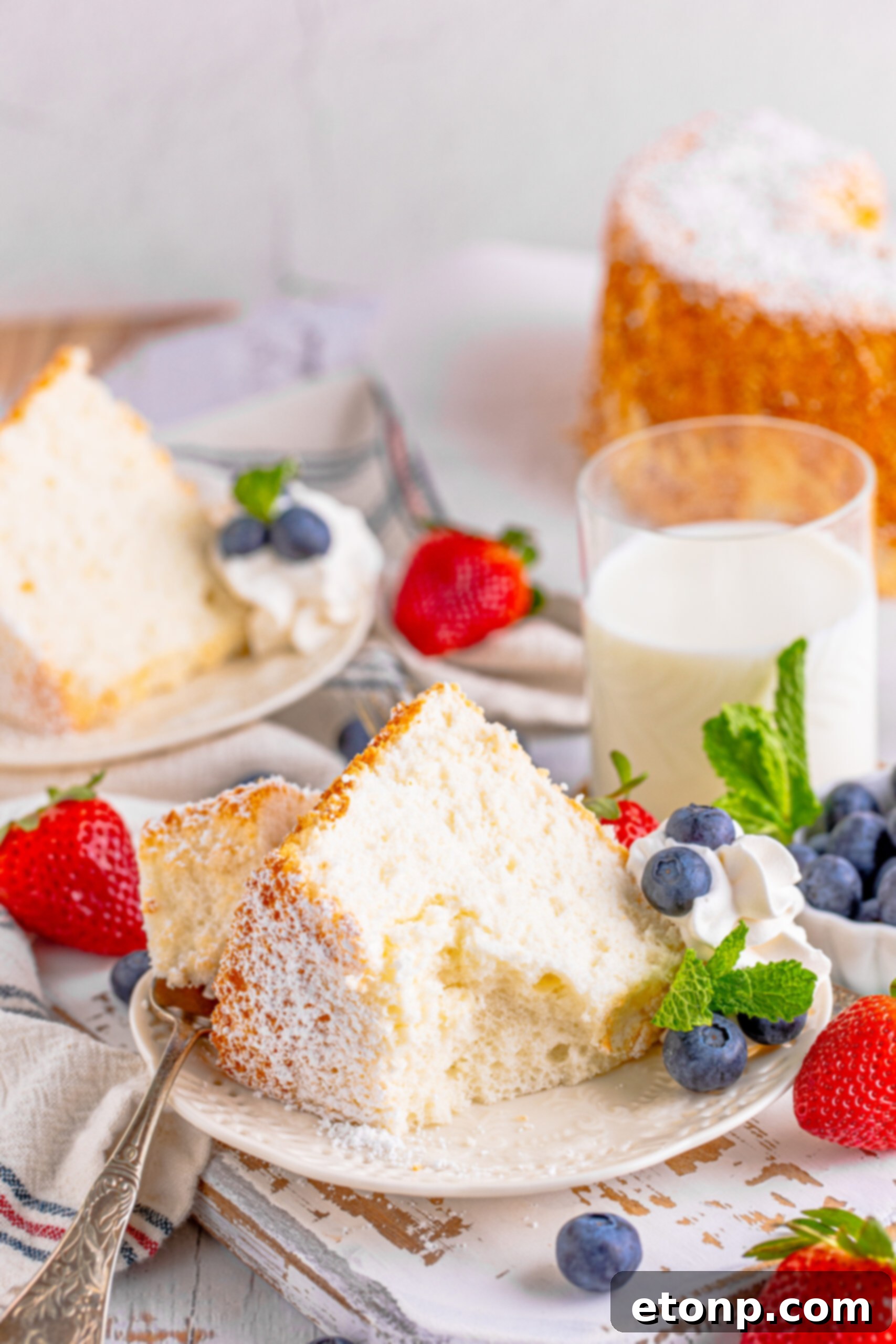 A piece of Angel Food Cake on a white plate with a bite removed on a silver fork with whipped cream, fresh berries, and mint