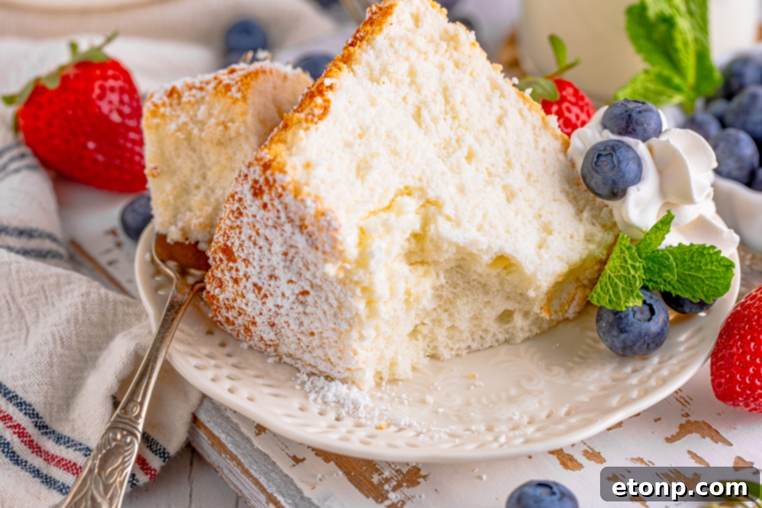 A piece of Angel Food Cake on a white plate with a bite removed on a silver fork with whipped cream, fresh berries, and mint