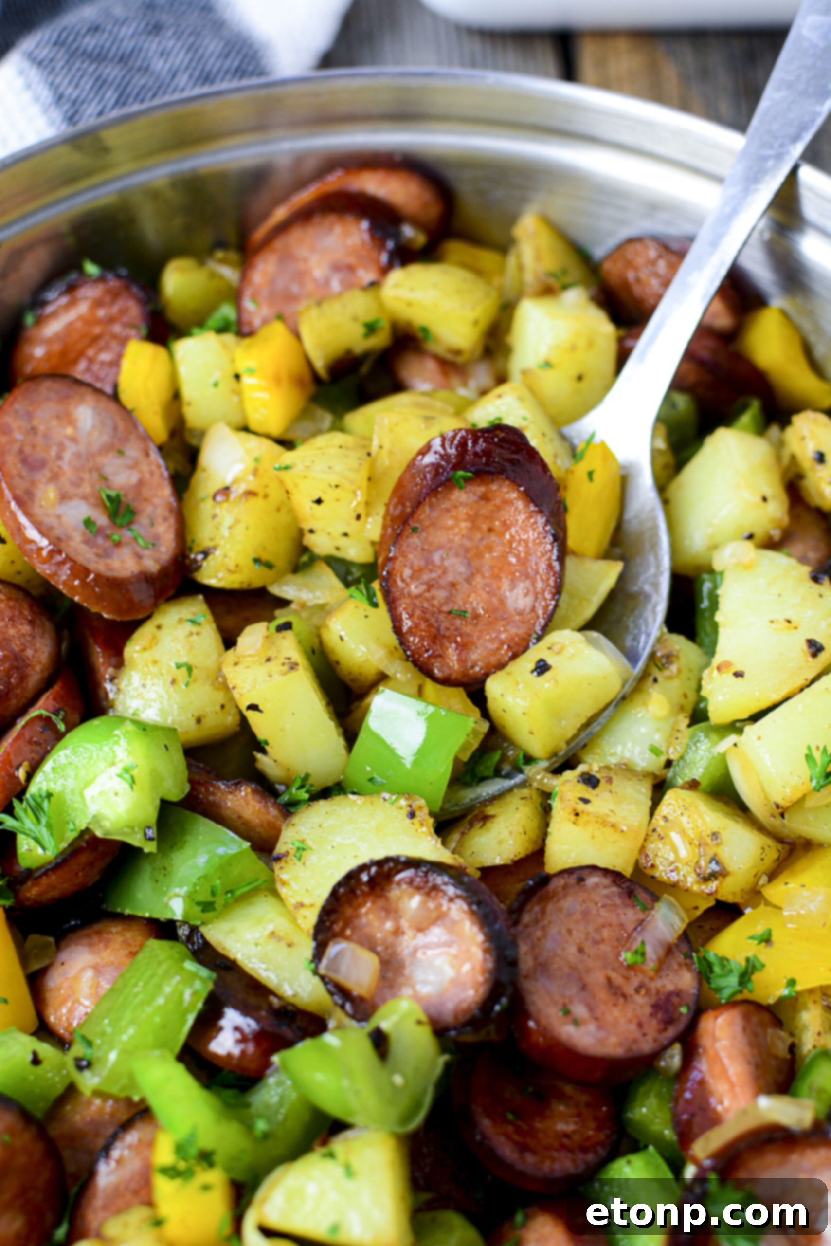 Overhead view of Sausage peppers and potatoes in a silver skillet with a spoon removing some for serving. 
