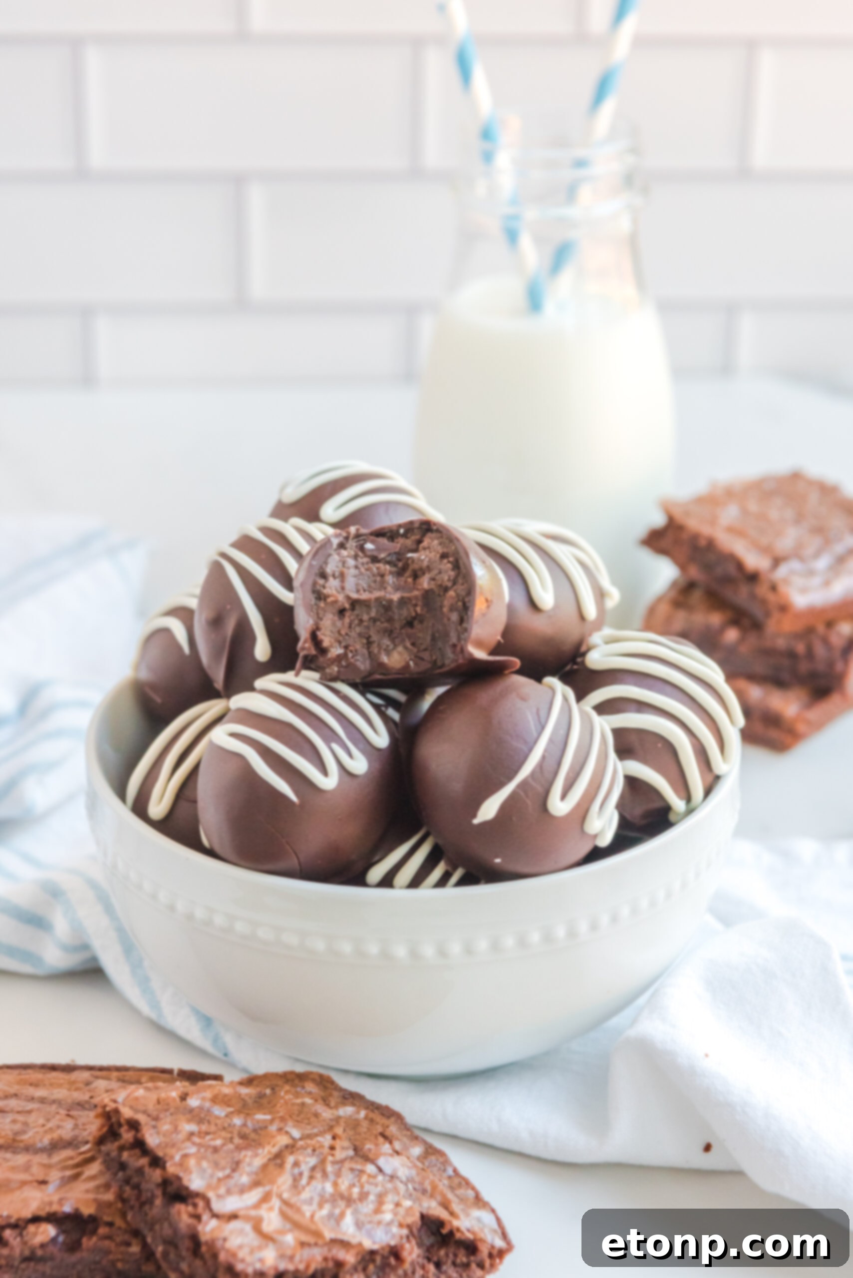 Decadent Brownie Truffles 15 A cozy scene featuring stacked Brownie Truffles in a white bowl, accompanied by brownies and a glass of milk with a charming blue and white straw in the background, inviting a moment of sweet indulgence.