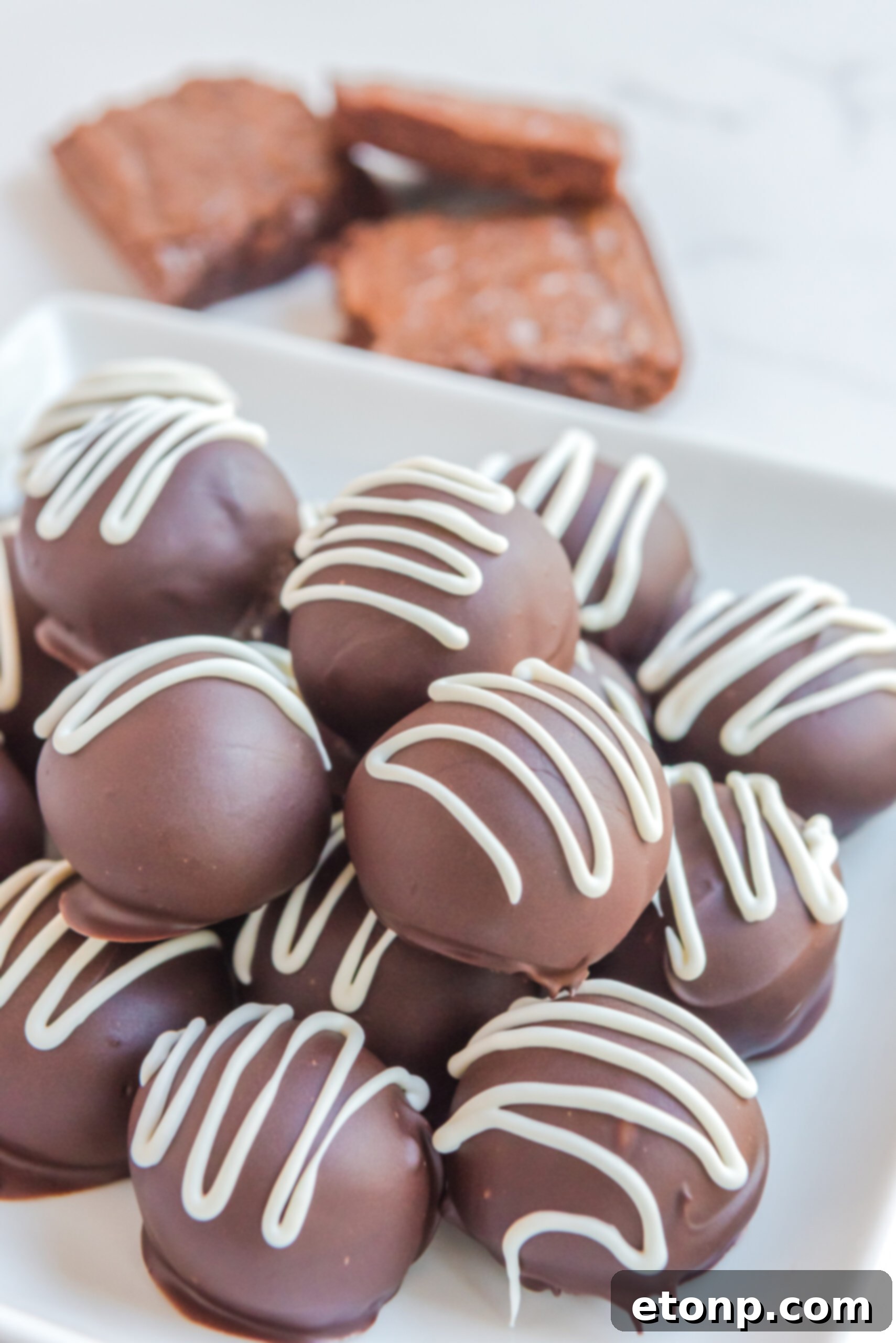 Decadent Brownie Truffles 3 A pristine white plate showcases beautifully arranged Brownie Bite Truffles, with a stack of brownies subtly blurred in the background, hinting at their origin.