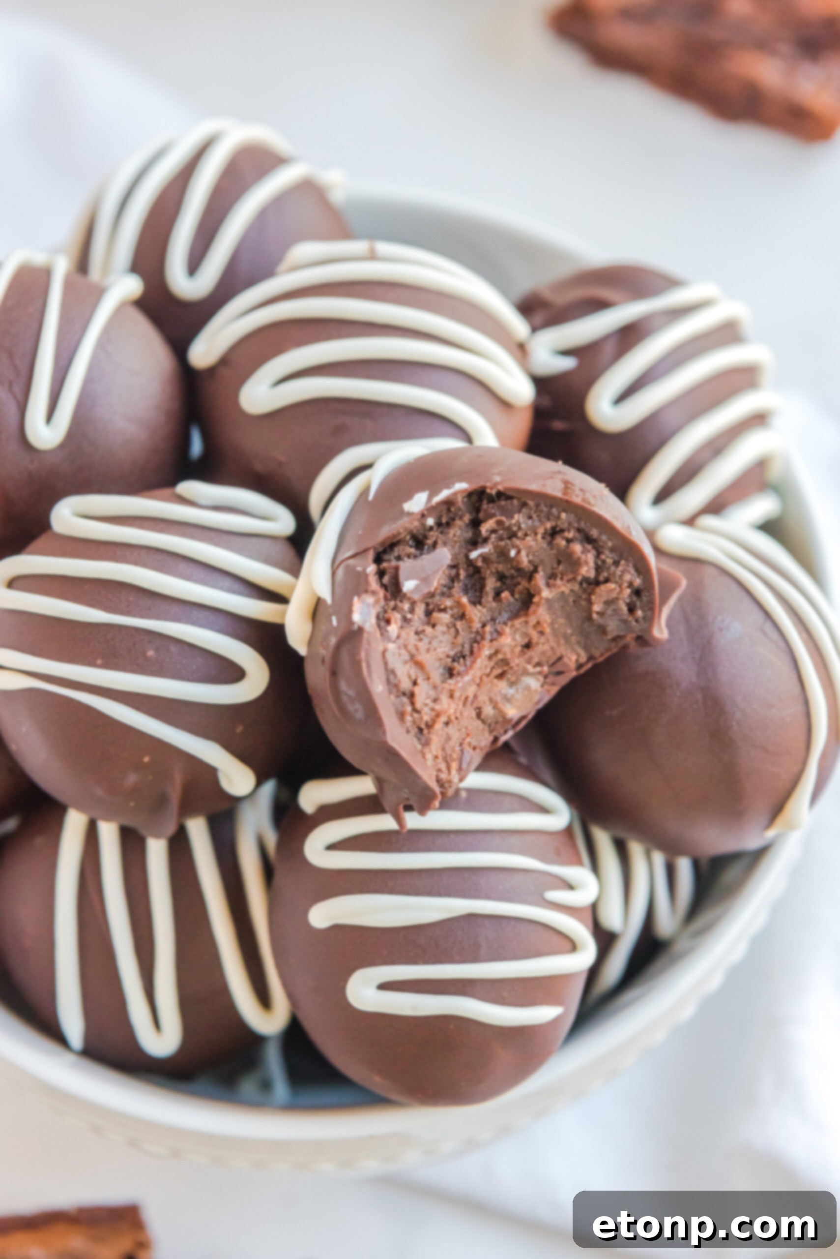 Decadent Brownie Truffles 5 Exquisite Brownie Bite Truffles artfully drizzled with delicate white chocolate, presented in a crisp white bowl, with one truffle invitingly bitten.
