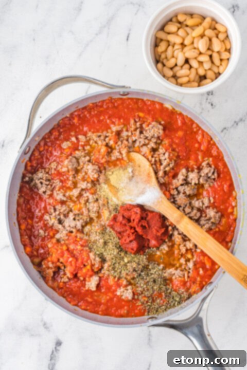 Cooked penne pasta being stirred into the rich meat and bean sauce.
