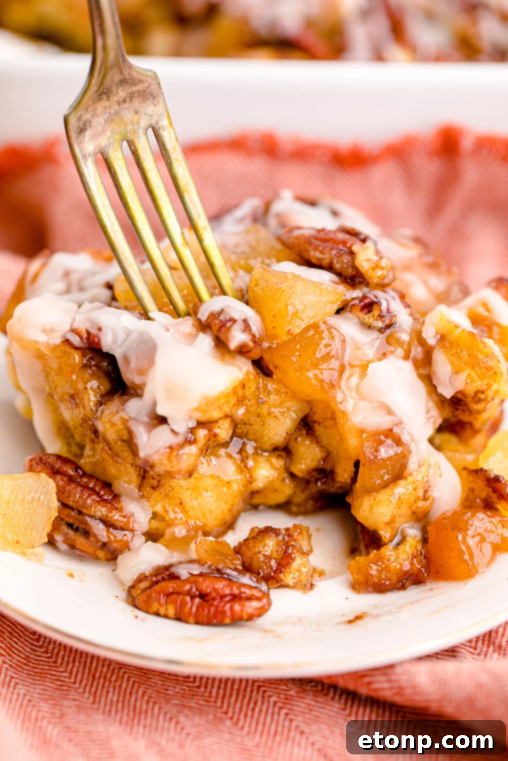 A golden fork lifts a bite-sized piece of Cinnamon Roll Casserole from a white plate, showcasing its gooey texture and sweet icing.