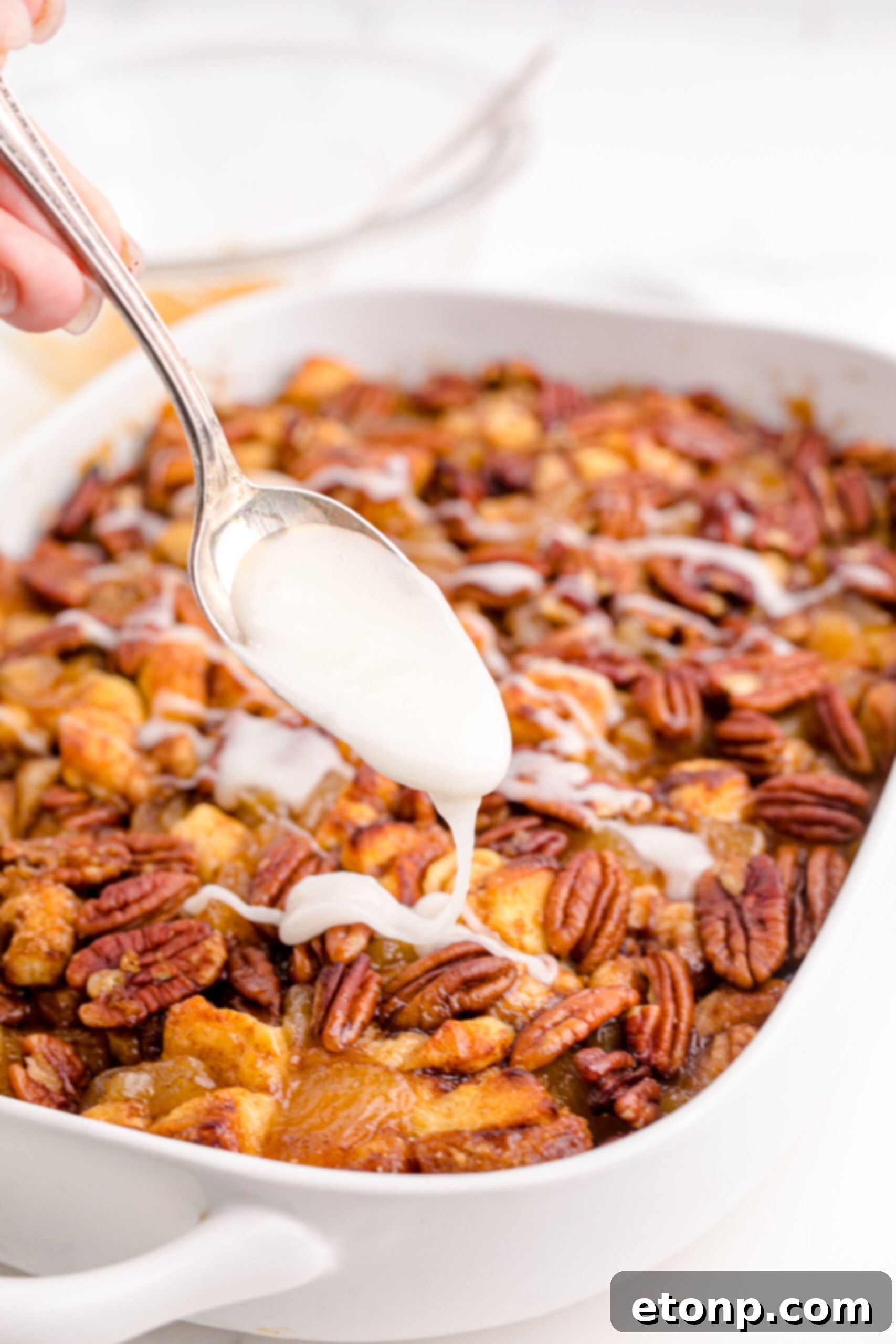 A spoon drizzles warm icing over the baked Cinnamon Roll Casserole in a white baking dish, making it extra gooey and appealing.