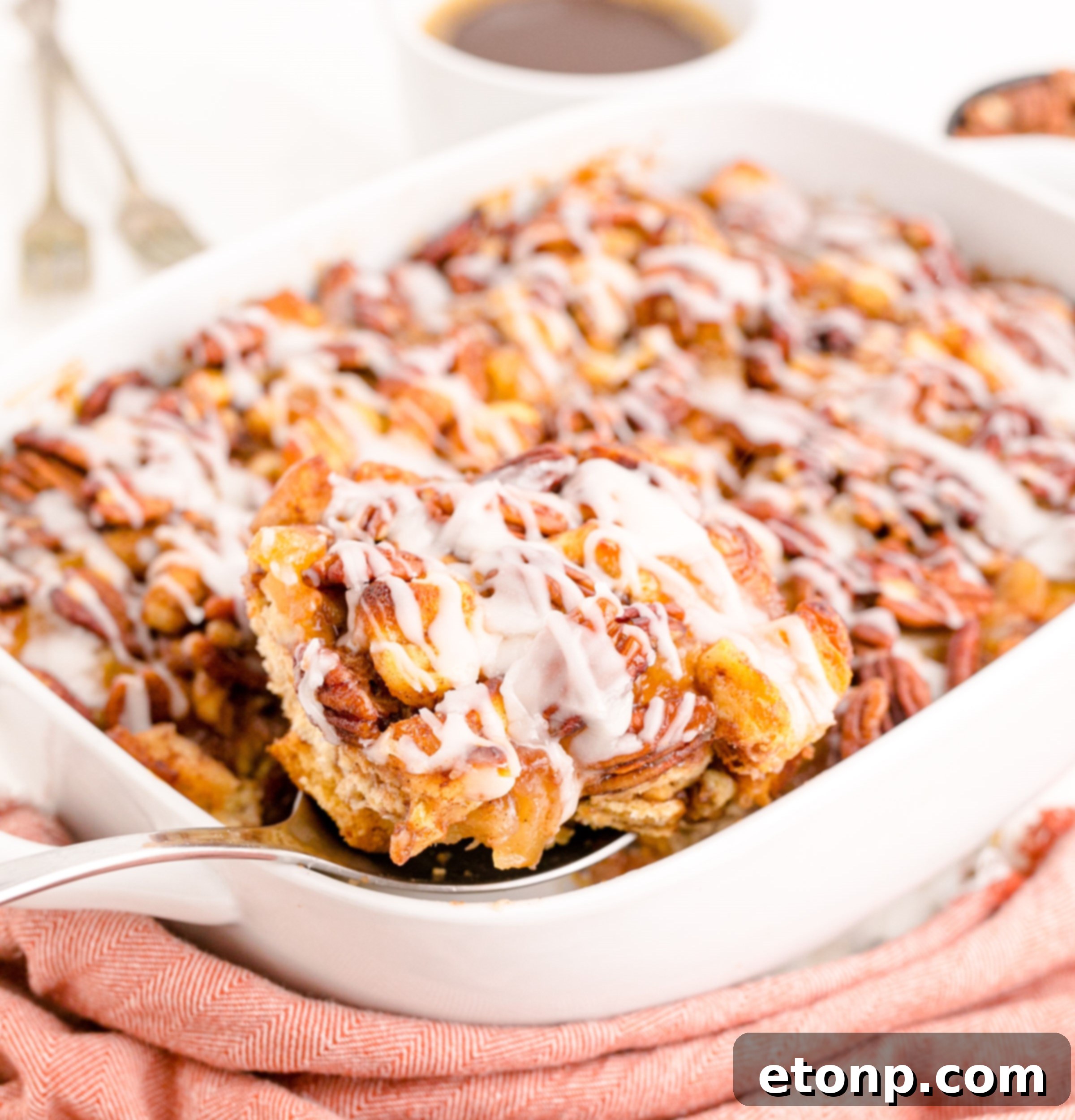 A silver spoon scoops a generous serving of Cinnamon Roll Casserole from a pristine white baking dish, revealing layers of cinnamon-swirled goodness.