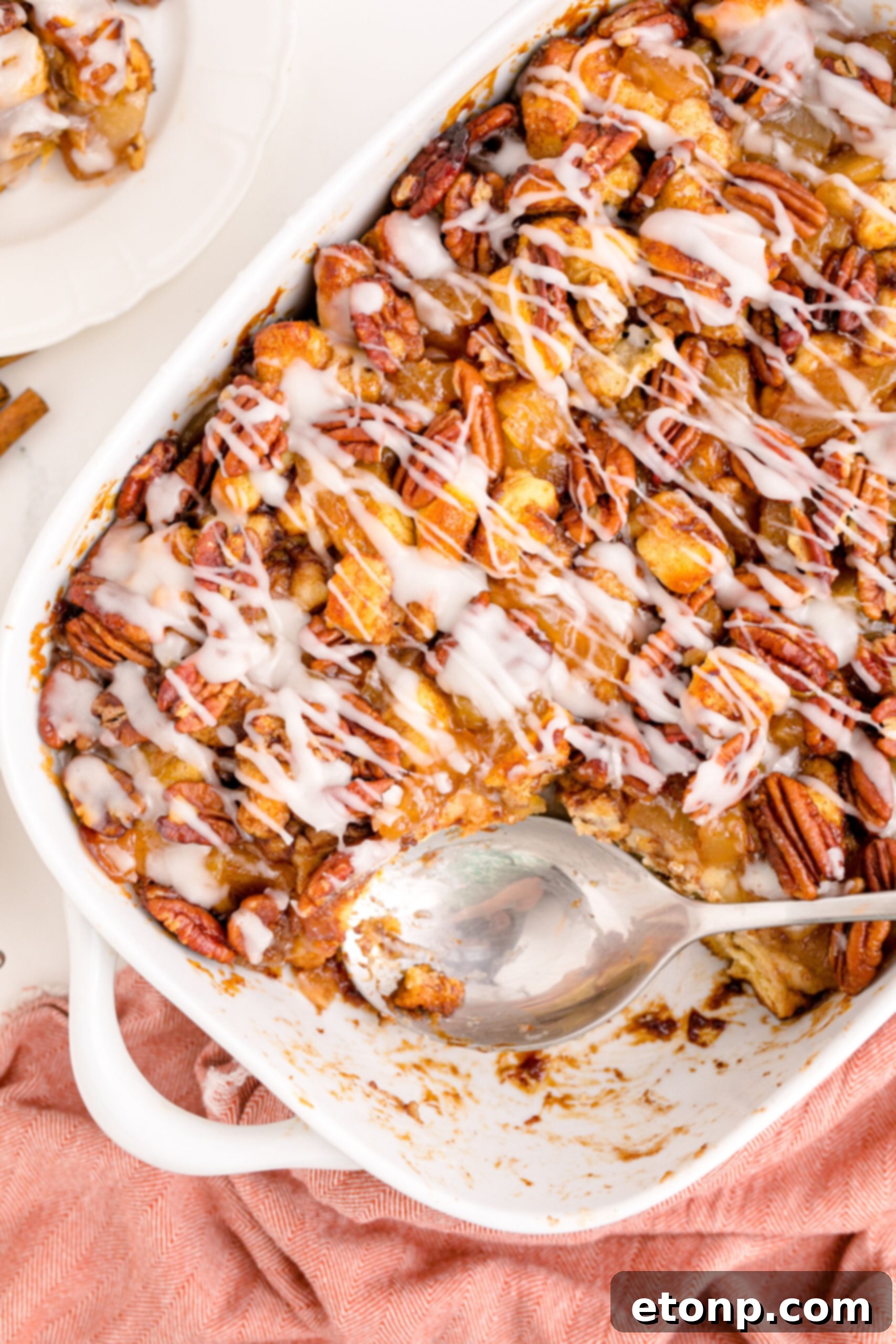 An overhead shot shows a silver spoon carefully lifting a portion of the baked Cinnamon Roll Casserole from a white baking dish, highlighting its golden-brown topping and rich textures.