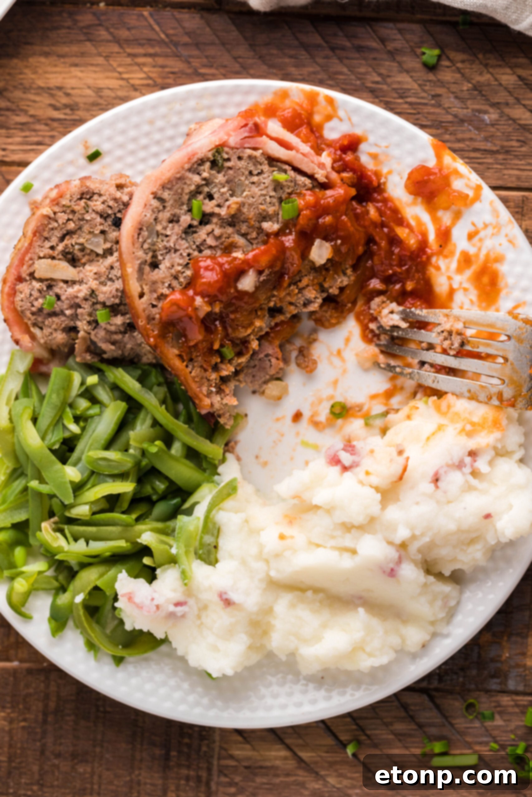 Bacon-Cloaked Meatloaf 17 Overhead view of a slice of Bacon Wrapped Meatloaf, cut in half by a silver fork, on a white dish served with mashed potatoes and green beans.