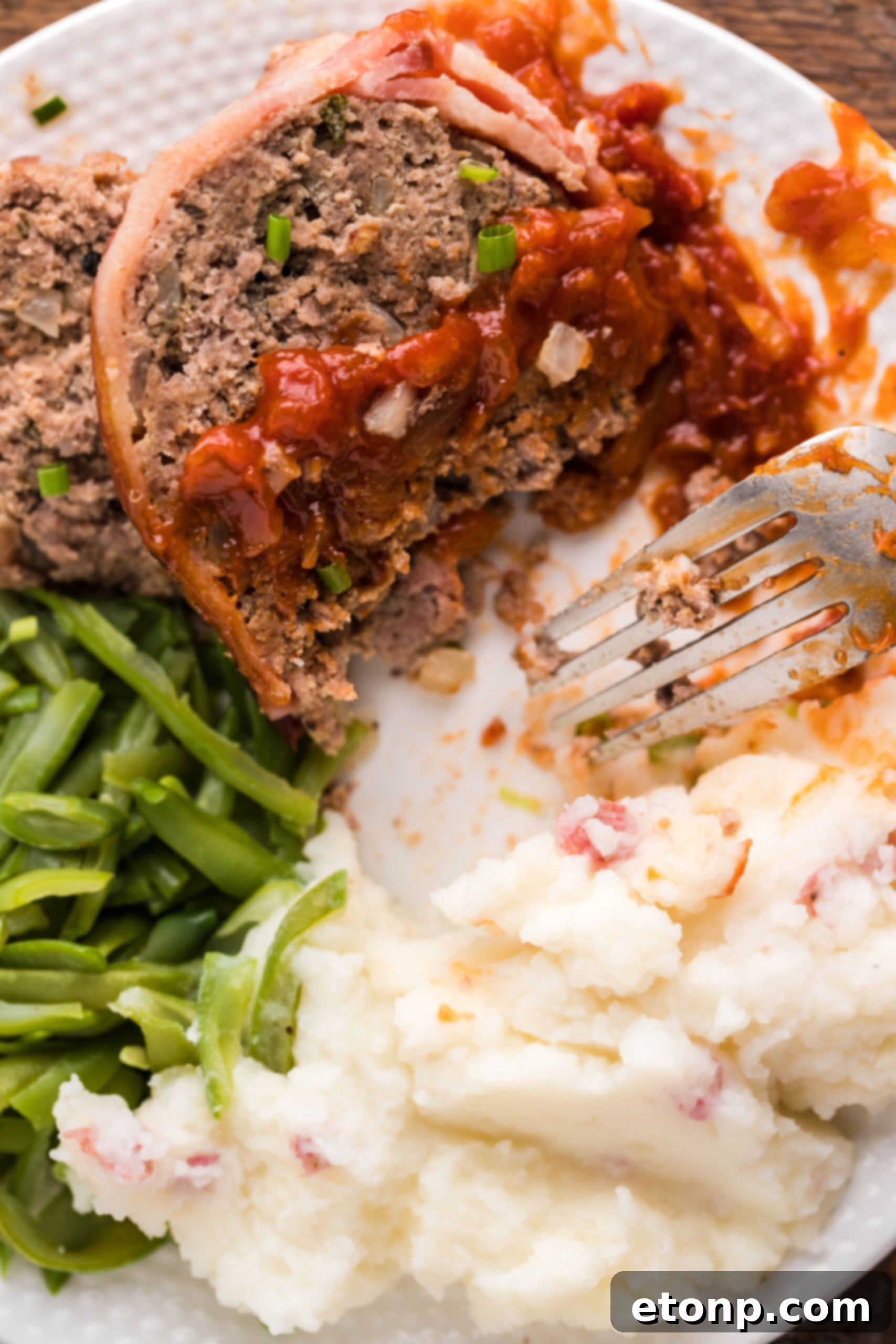 Bacon-Cloaked Meatloaf 6 Close up view of a slice of Bacon Wrapped Meatloaf, cut in half by a silver fork, on a white dish served with mashed potatoes and green beans.