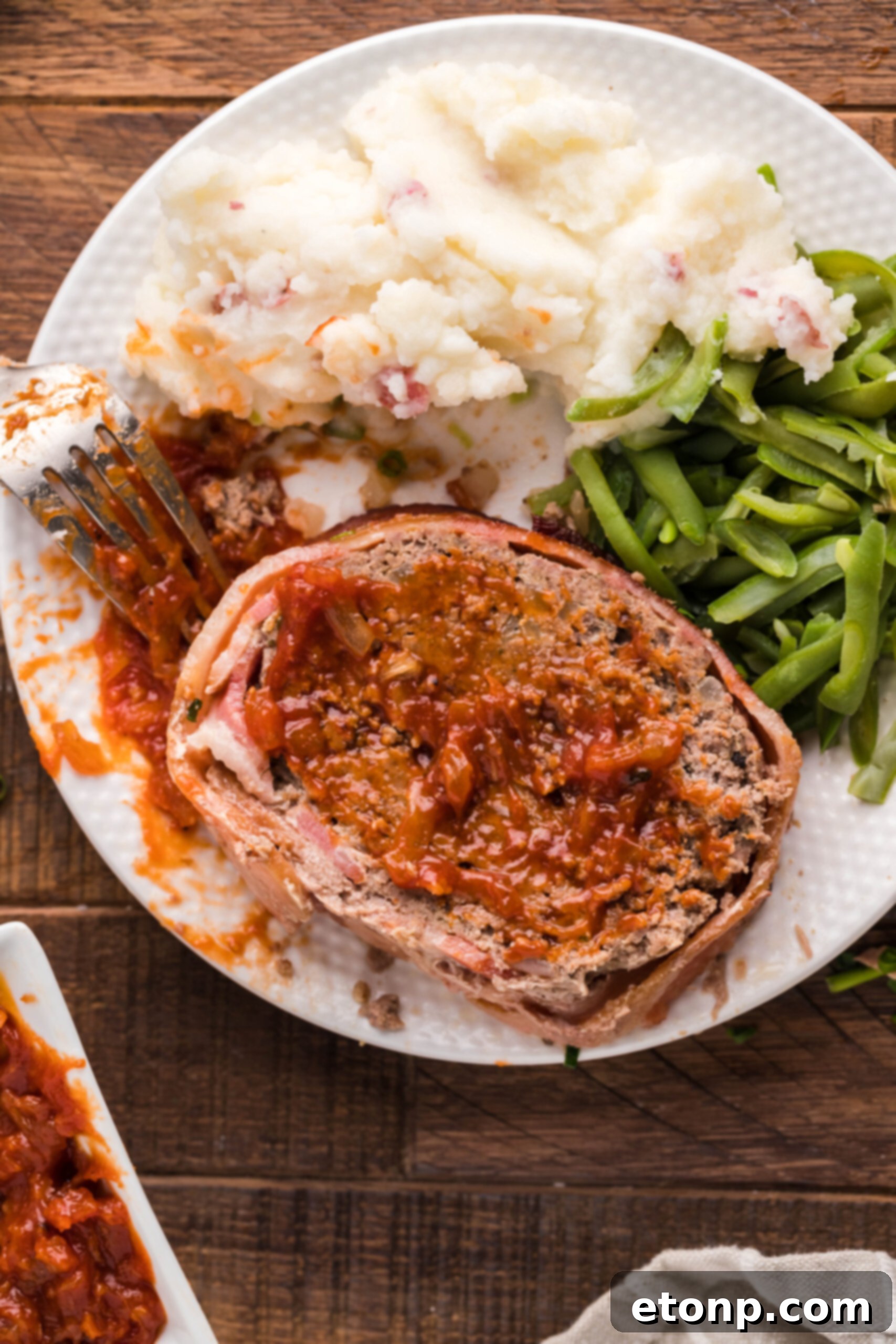 Bacon-Cloaked Meatloaf 8 Overhead view of Bacon Wrapped Meatloaf on a white dish served with mashed potatoes and green beans.