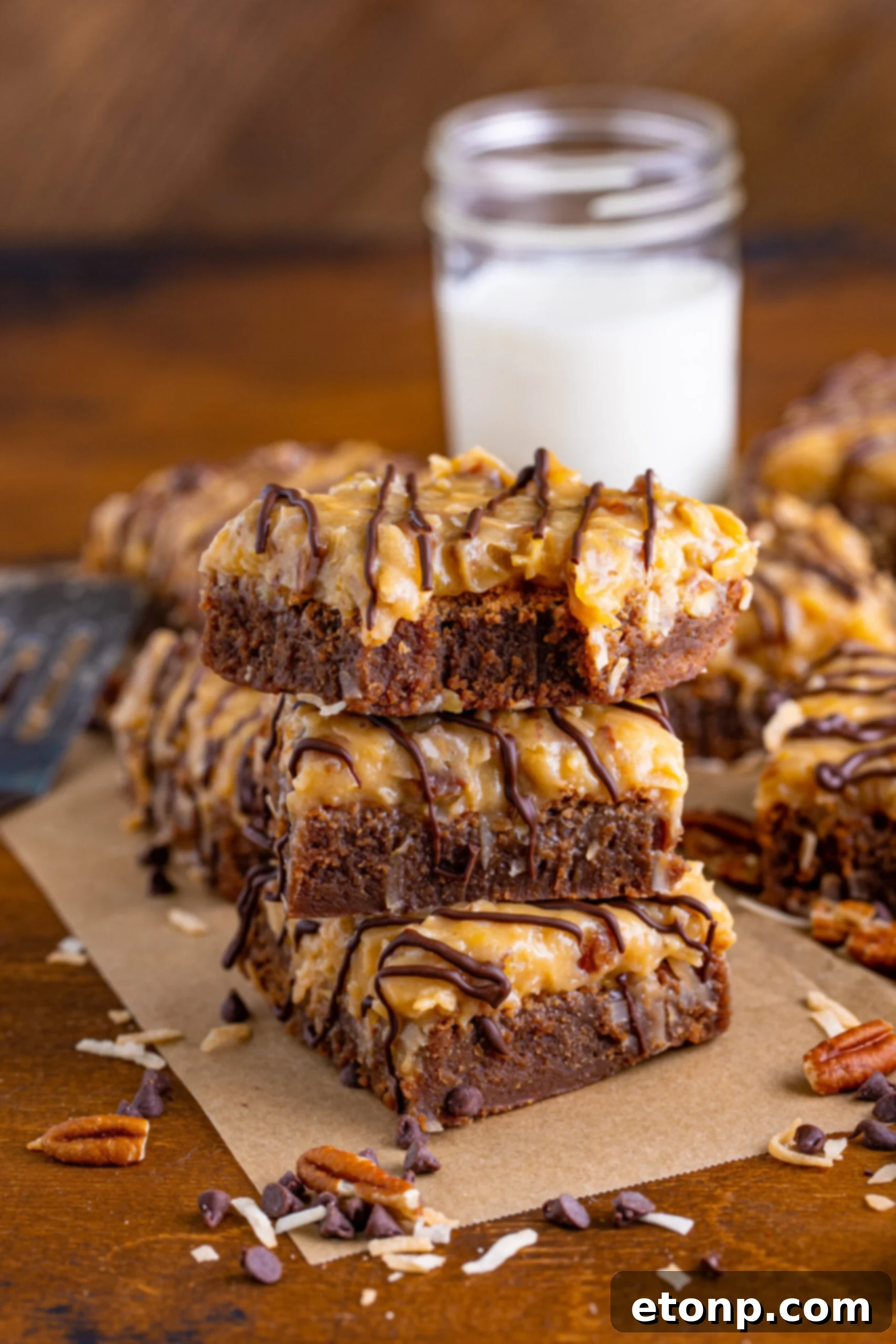 Front view of three stacked German Chocolate Brownies on parchment paper, with a bite removed from the top brownie, showcasing the rich texture and coconut-pecan topping.