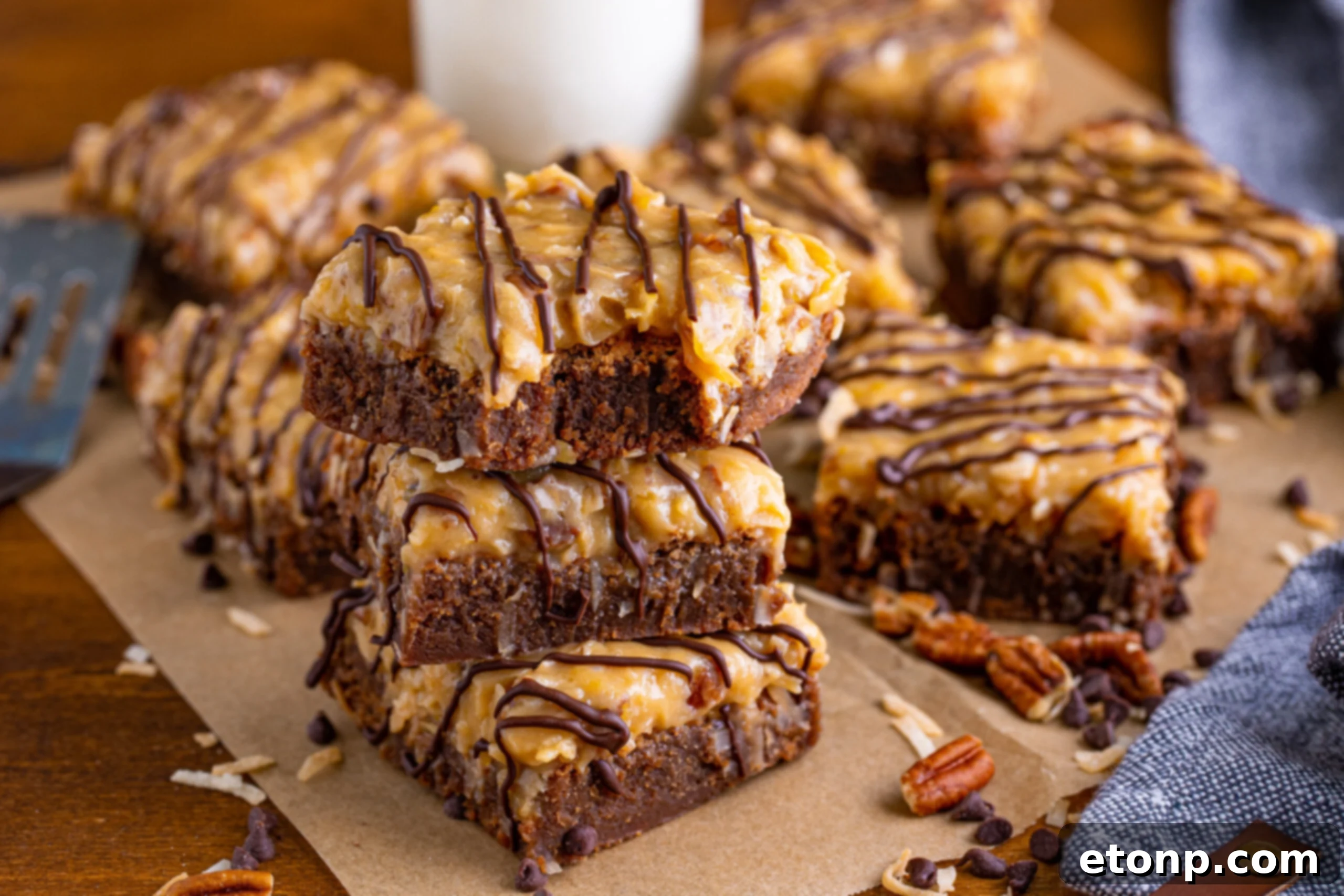 Three German Chocolate Brownies stacked on a wood table with a bite missing from the top one, artistically surrounded by chopped pecans and chocolate chips.