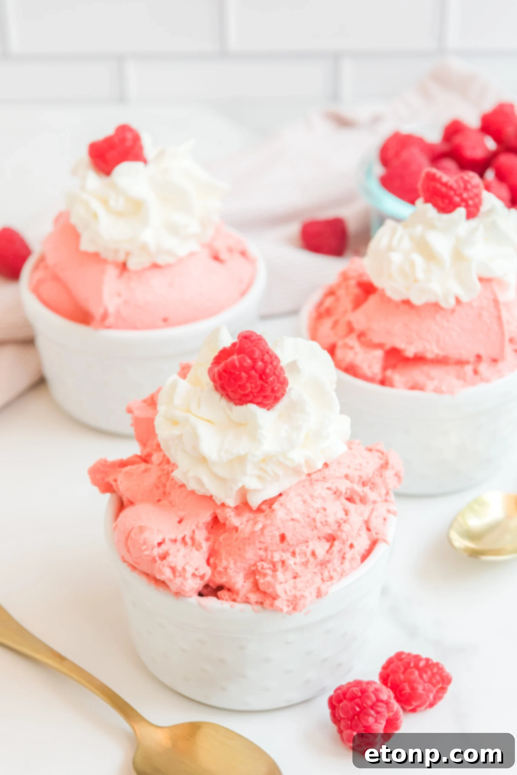 Front view of Raspberry Jello Fluff in three white dishes garnished with whipped cream and fresh raspberries. The dessert is a light pink color, smooth and fluffy, with a few fresh raspberries and a dollop of white whipped topping on each.