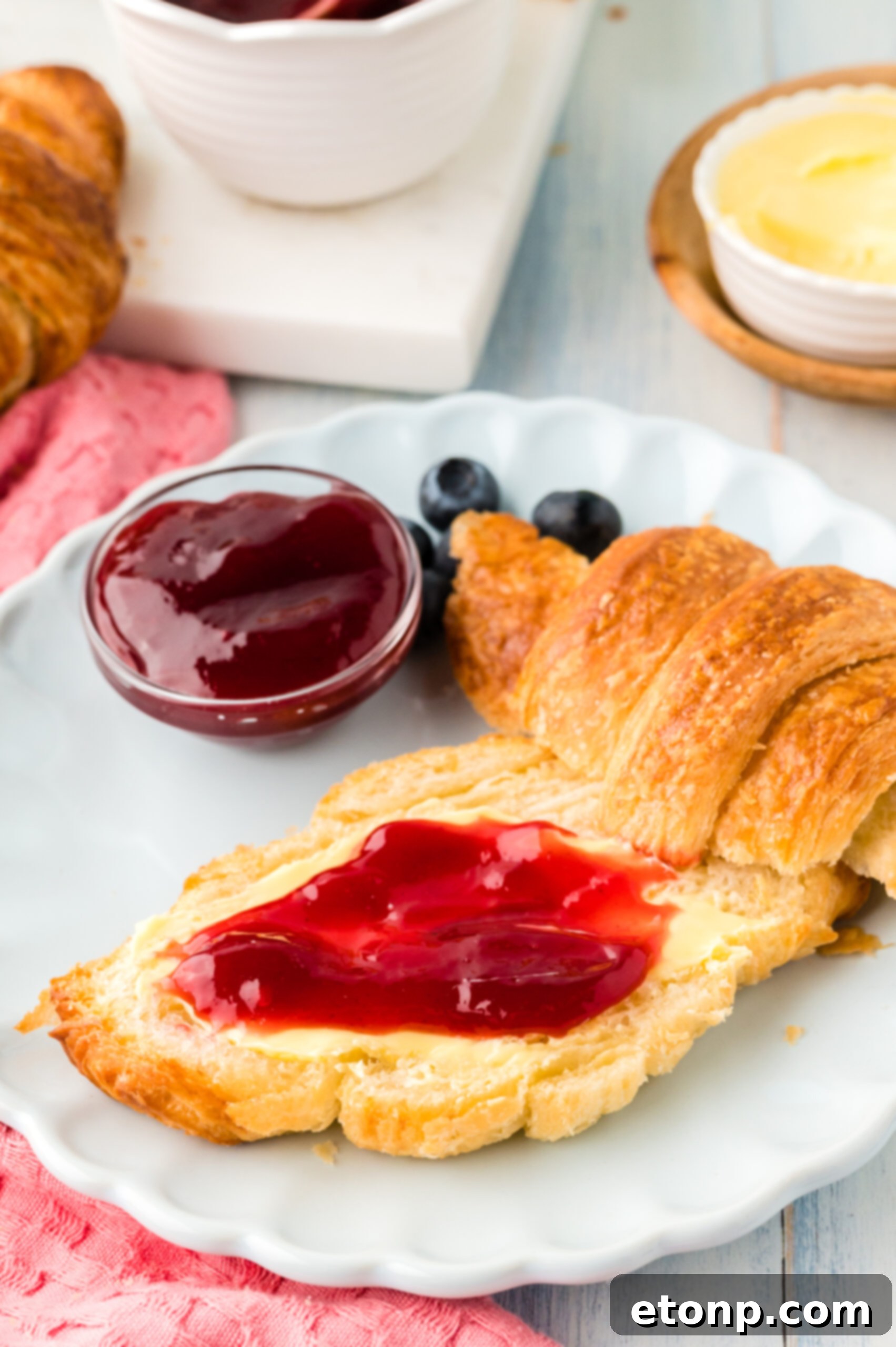 Front view of Homemade Croissants cut in half with jam spread on the bottom half, served on a white dish with jam and fresh blueberries.