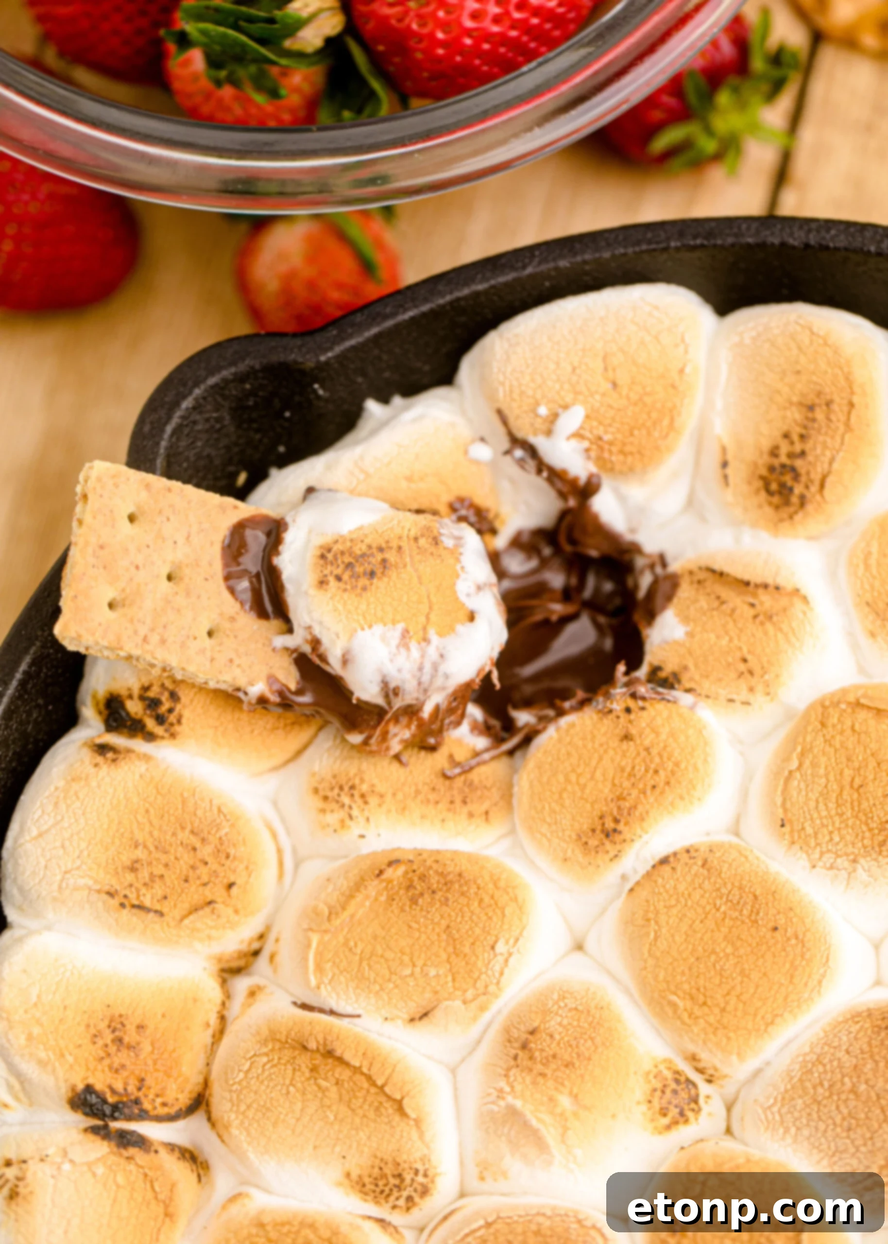 A close-up of a graham cracker scooping a generous portion of warm S'mores Dip from a hot skillet, showcasing the melted chocolate and stretched marshmallow.