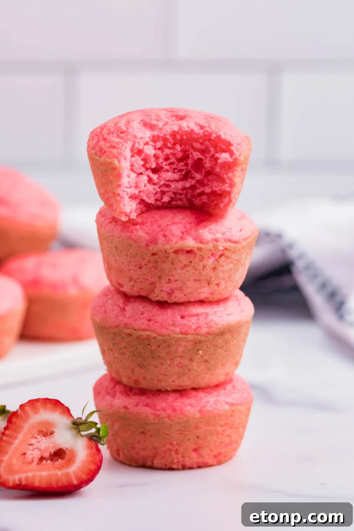 Four delightful Weight Watchers Strawberry Muffins stacked artfully, with a bite taken from the top one, showcasing its tender, fruit-studded interior.