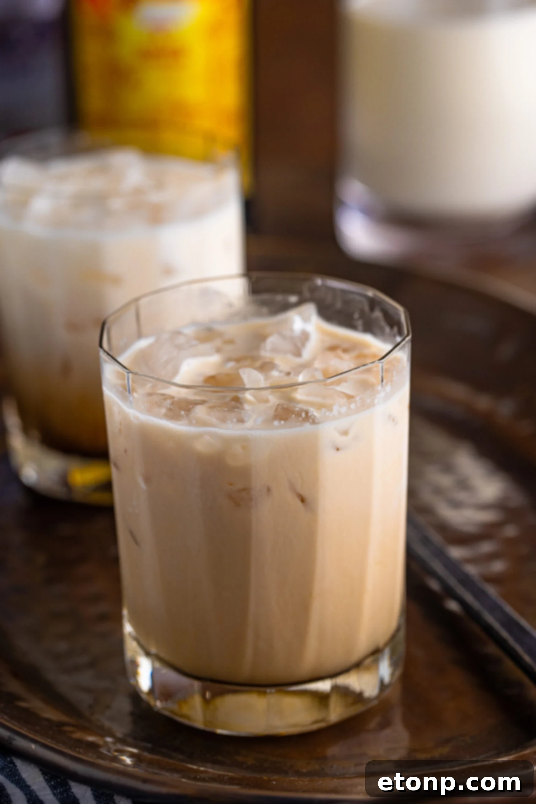 White Russian in a clear glass sitting on a dark metal serving tray. The cocktail is perfectly layered with white cream on top and a dark coffee liqueur and vodka base.