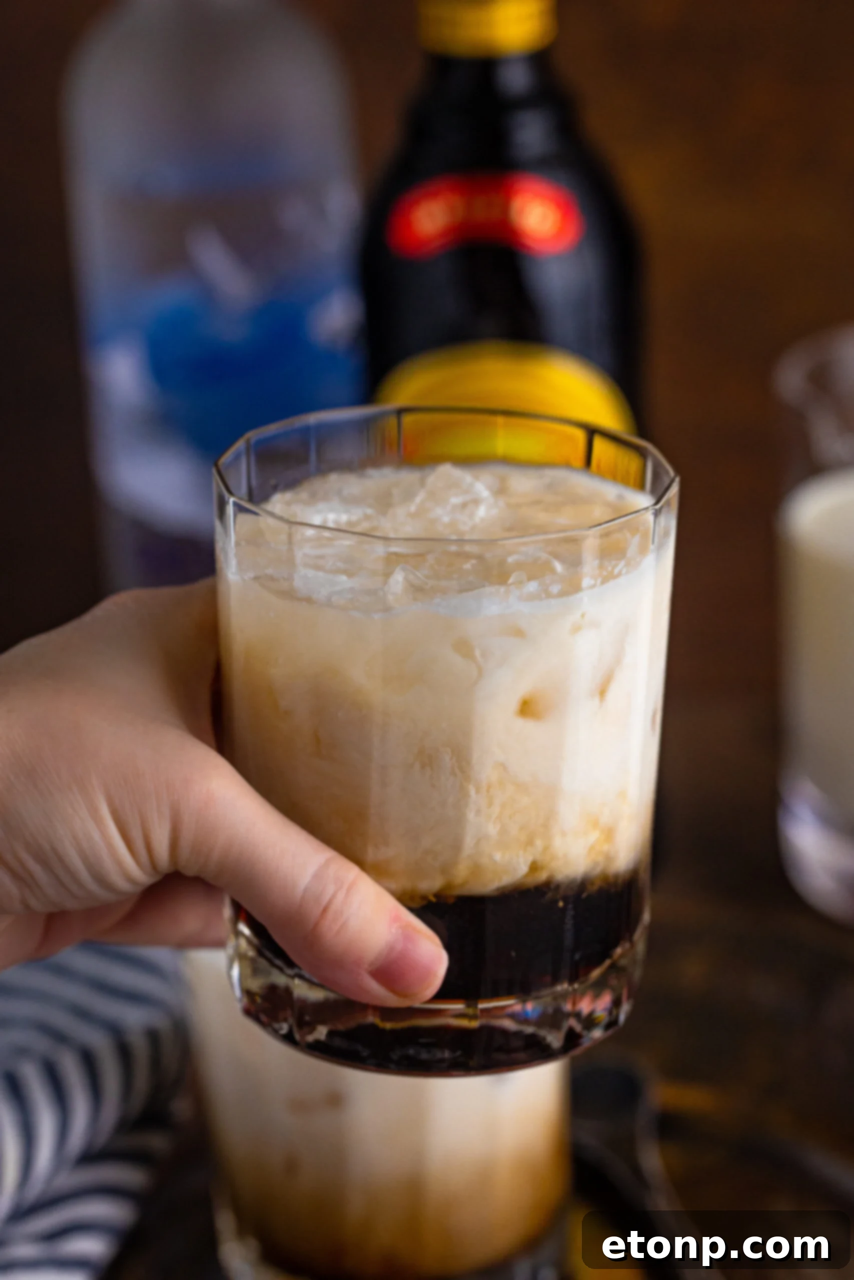 A hand holding a White Russian drink in a clear glass, showing the drink's creamy texture and appealing appearance.