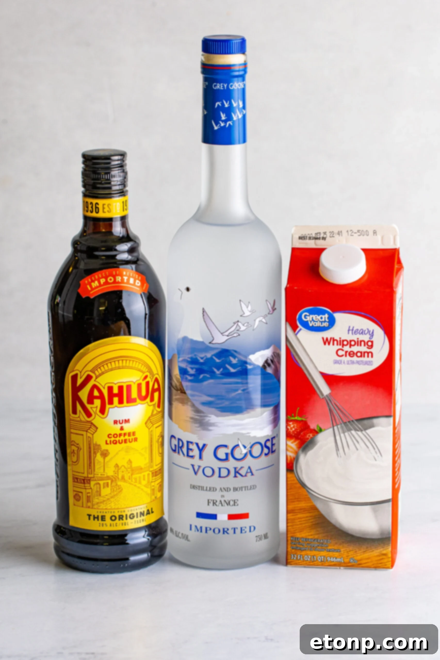 All essential ingredients needed for a White Russian cocktail: a bottle of vodka, a bottle of Kahlúa, and a carton of heavy cream, arranged neatly.