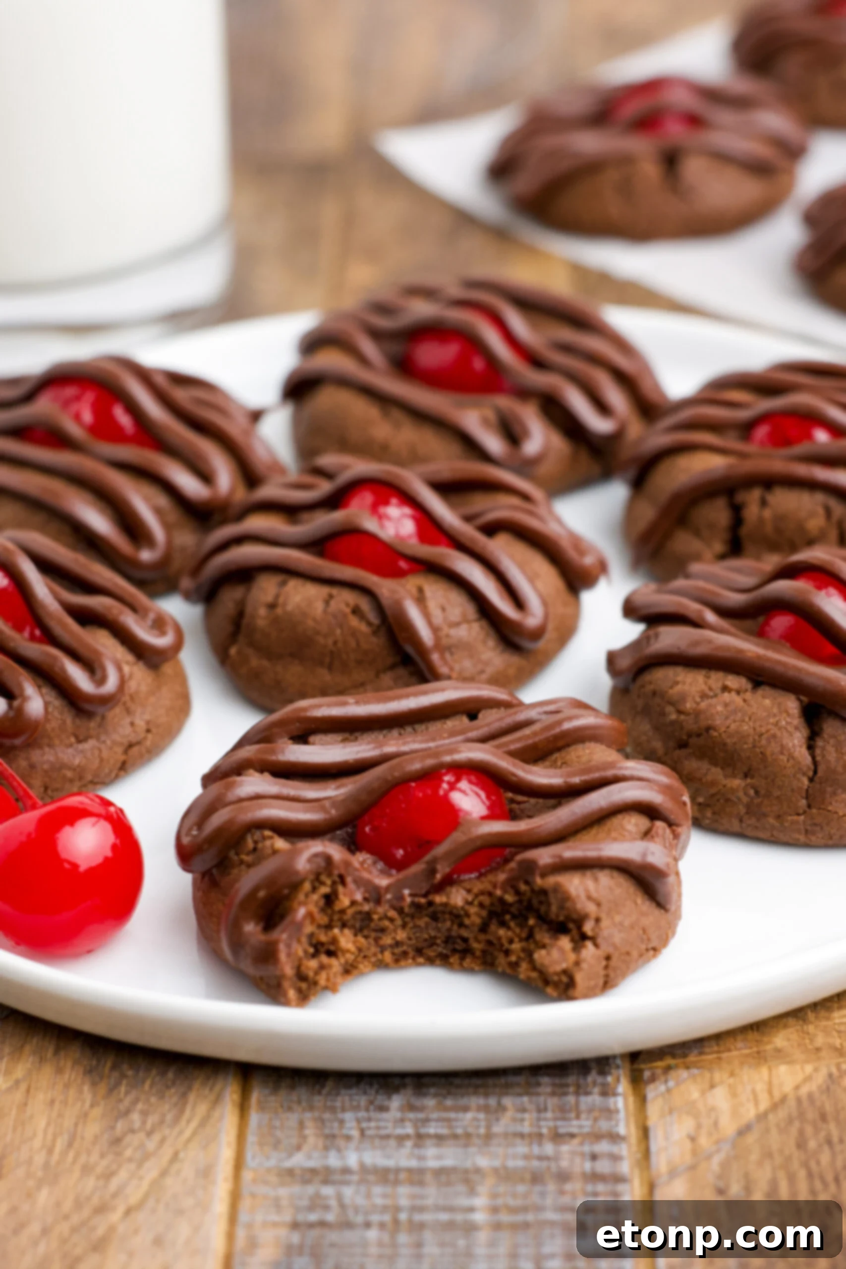 Close-up of freshly baked Chocolate Cherry Cookies on a pristine white plate, with a cookie in the foreground showing a clean bite taken, highlighting the soft, moist crumb and the sweet maraschino cherry.