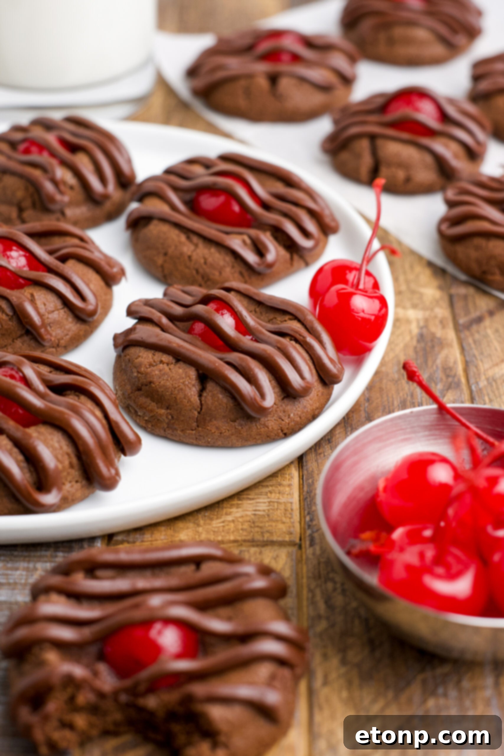 Beautifully arranged Chocolate Cherry Cookies on a white ceramic plate, with a scattering of bright red maraschino cherries in the background, emphasizing the key ingredient.