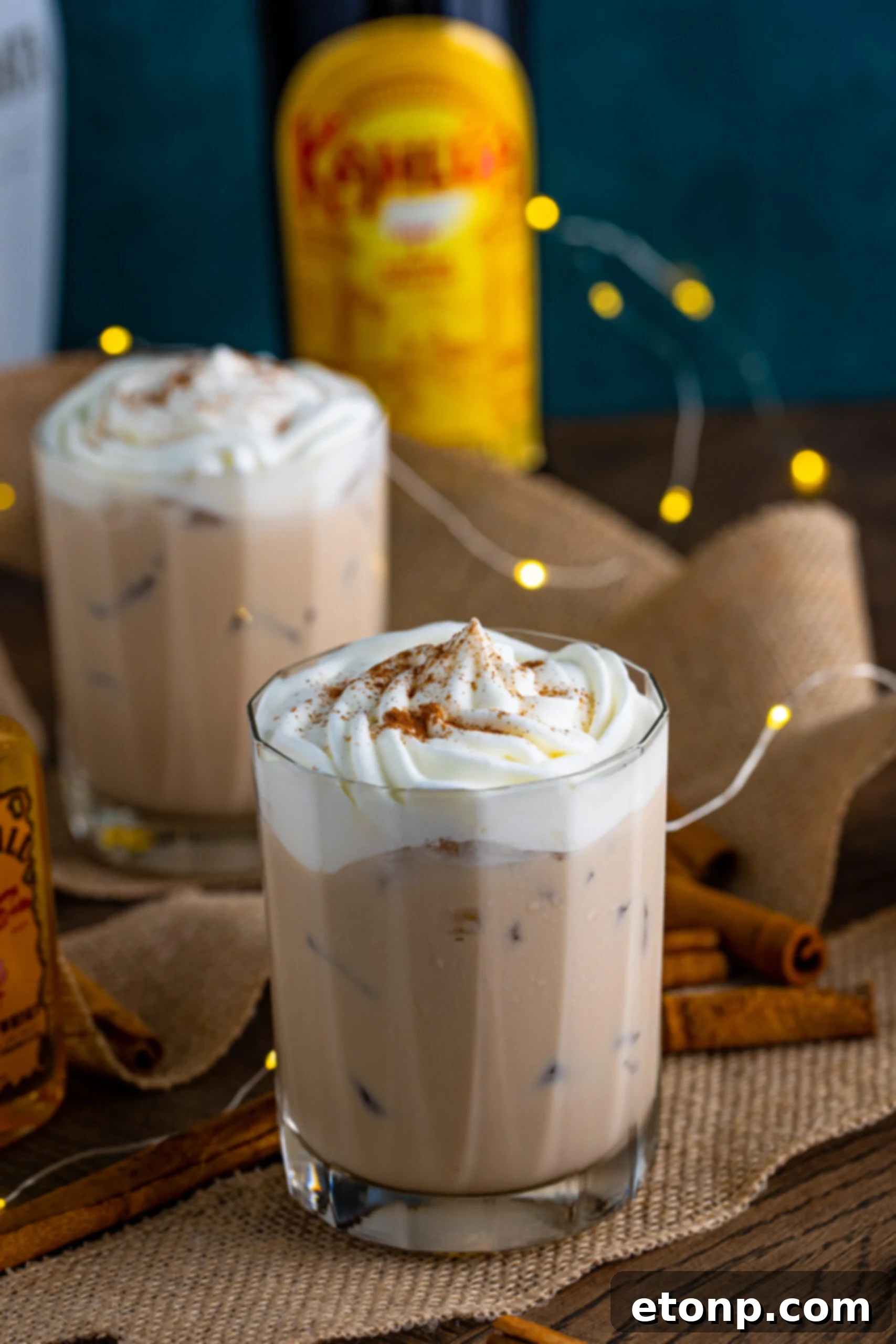 The cinnamon roll cocktail in a clear glass topped with whipped cream and dusted with cinnamon on a dark wood table surrounded by cinnamon sticks