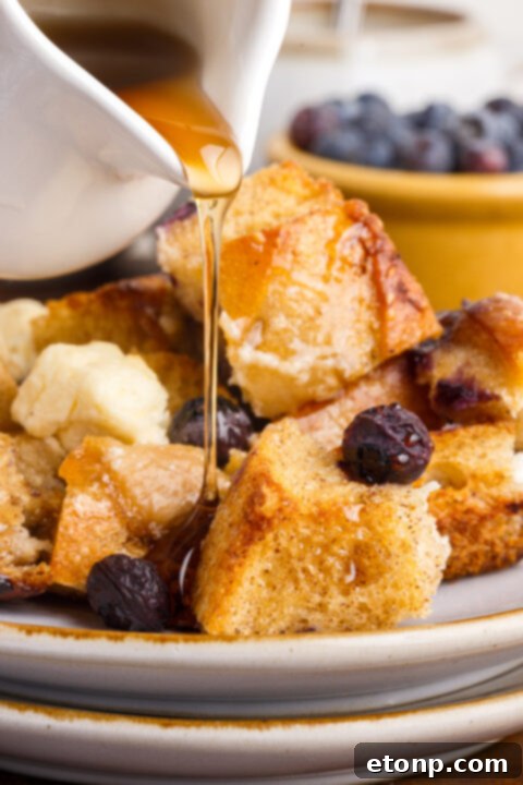 A beautifully presented serving of Blueberry French Toast Casserole, garnished with fresh blueberries and a dusting of powdered sugar, with maple syrup in a pitcher.