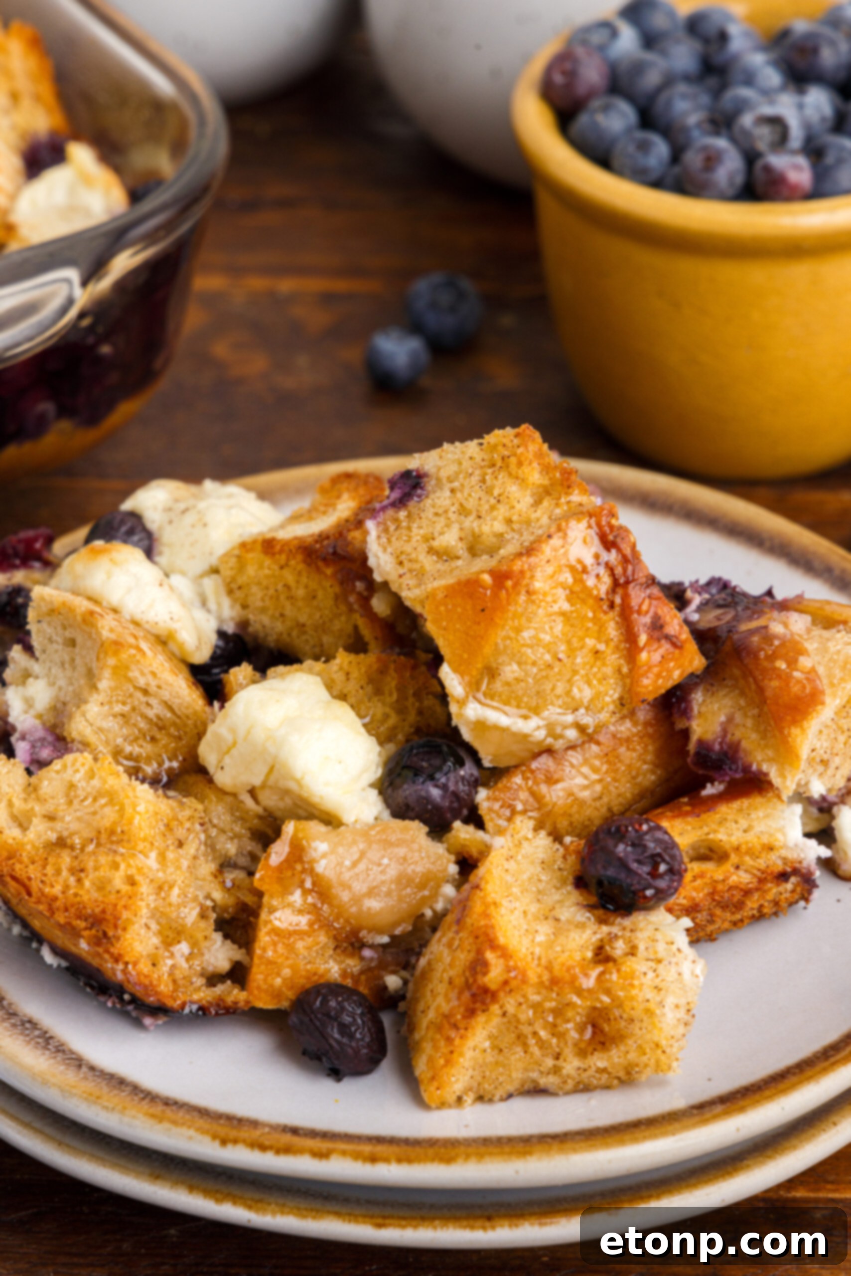 Close-up view of a single, perfectly golden-brown serving of Blueberry French Toast Casserole on a pristine white dish, showcasing the plump blueberries and fluffy texture.