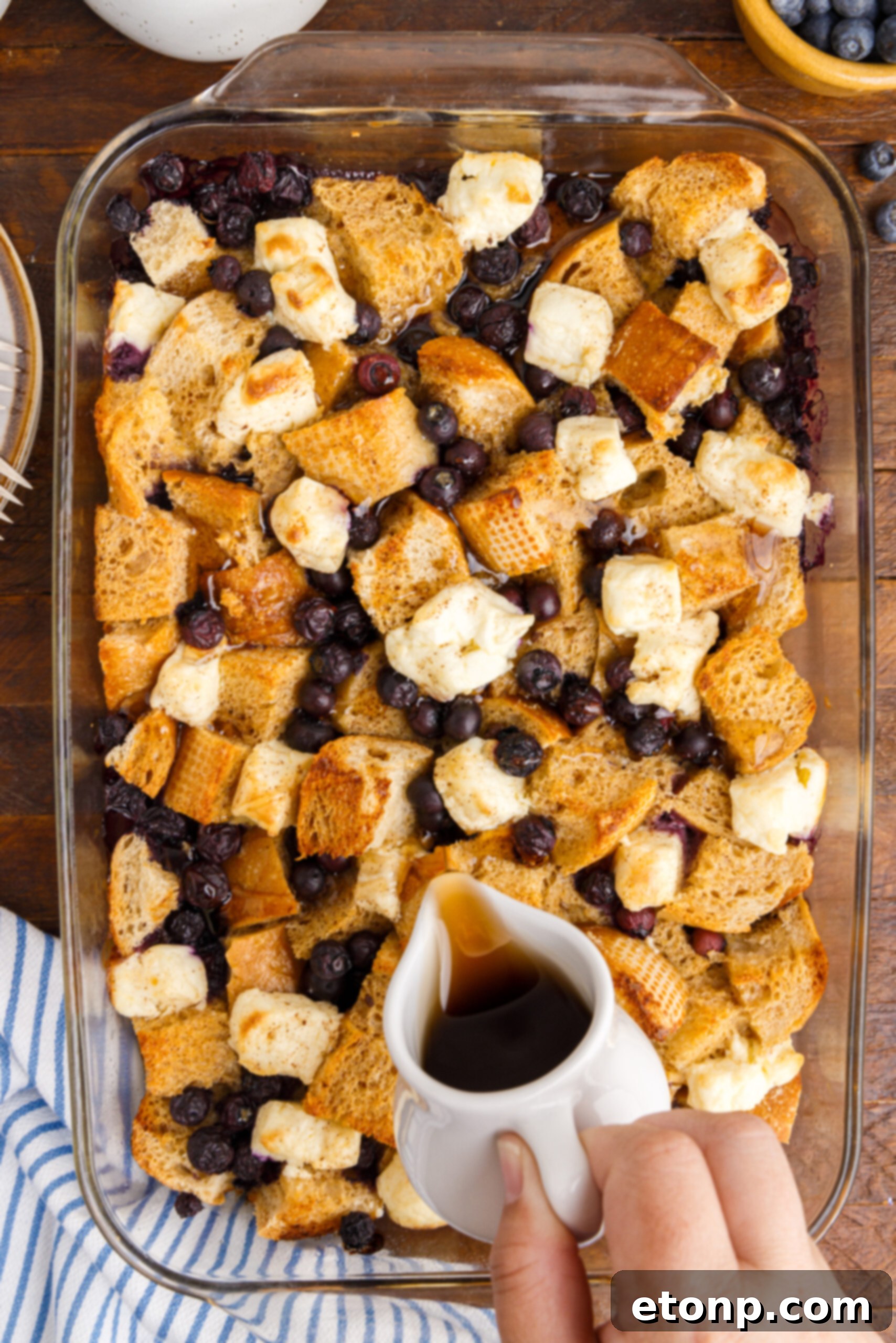 Overhead view of a clear baking dish filled with golden Blueberry French Toast Casserole, with warm syrup being drizzled over the top.