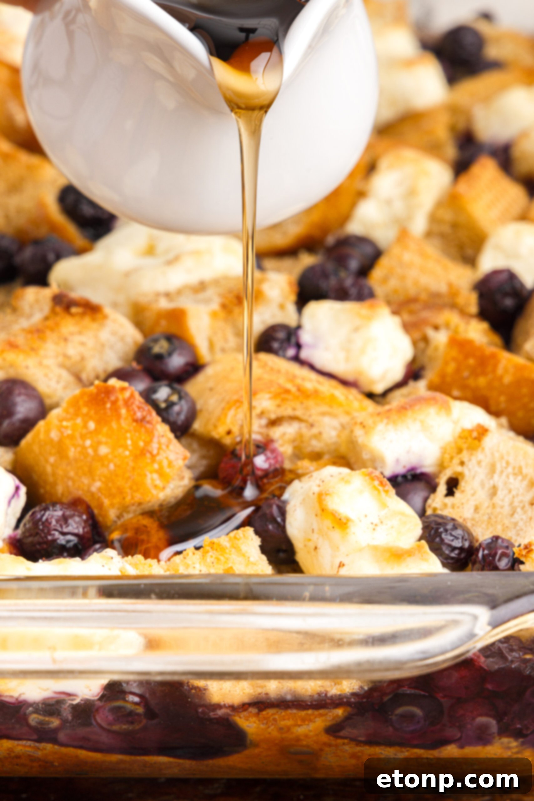 Detailed view of Blueberry French Toast Casserole in a clear glass baking dish, with a stream of maple syrup cascading over the top, highlighting its golden texture.