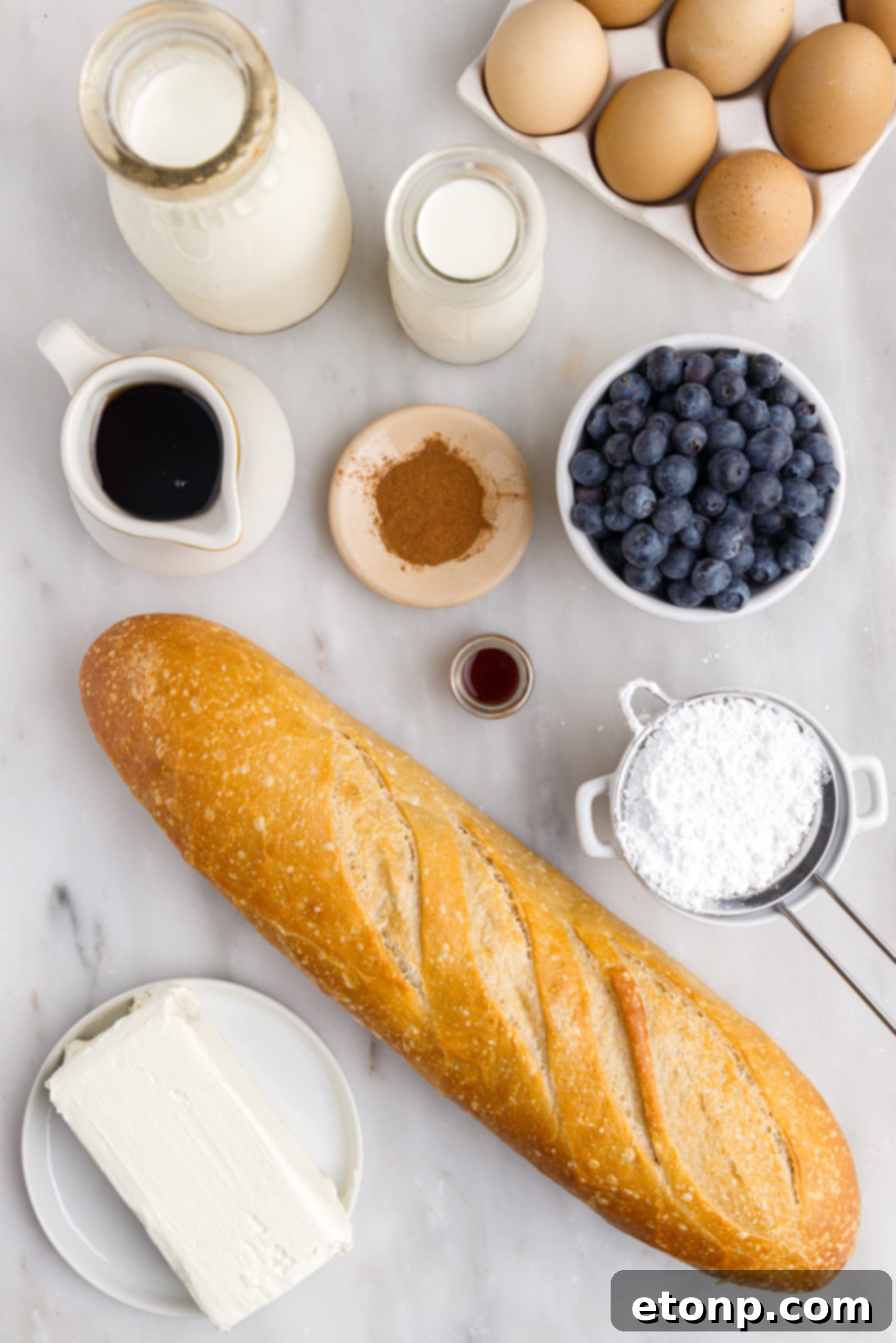 An inviting overhead shot displaying all the fresh ingredients needed for Blueberry French Toast Casserole laid out on a light surface: bread cubes, eggs, milk, heavy cream, maple syrup, cinnamon, vanilla extract, blueberries, and cream cheese.