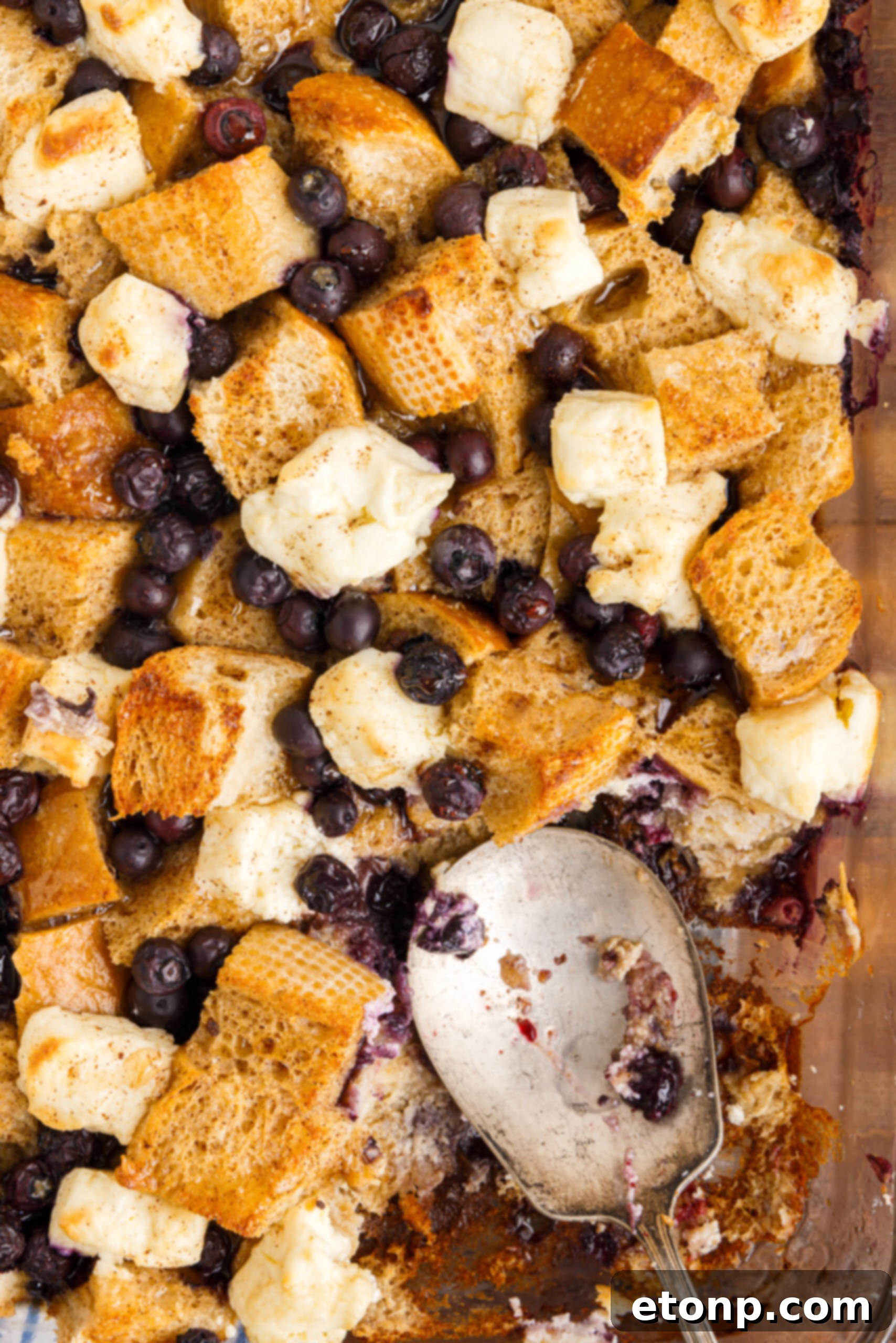Overhead shot of a clear glass baking dish with Blueberry French Toast Casserole, a portion elegantly removed with a silver serving spoon, revealing the soft interior and melted cream cheese.