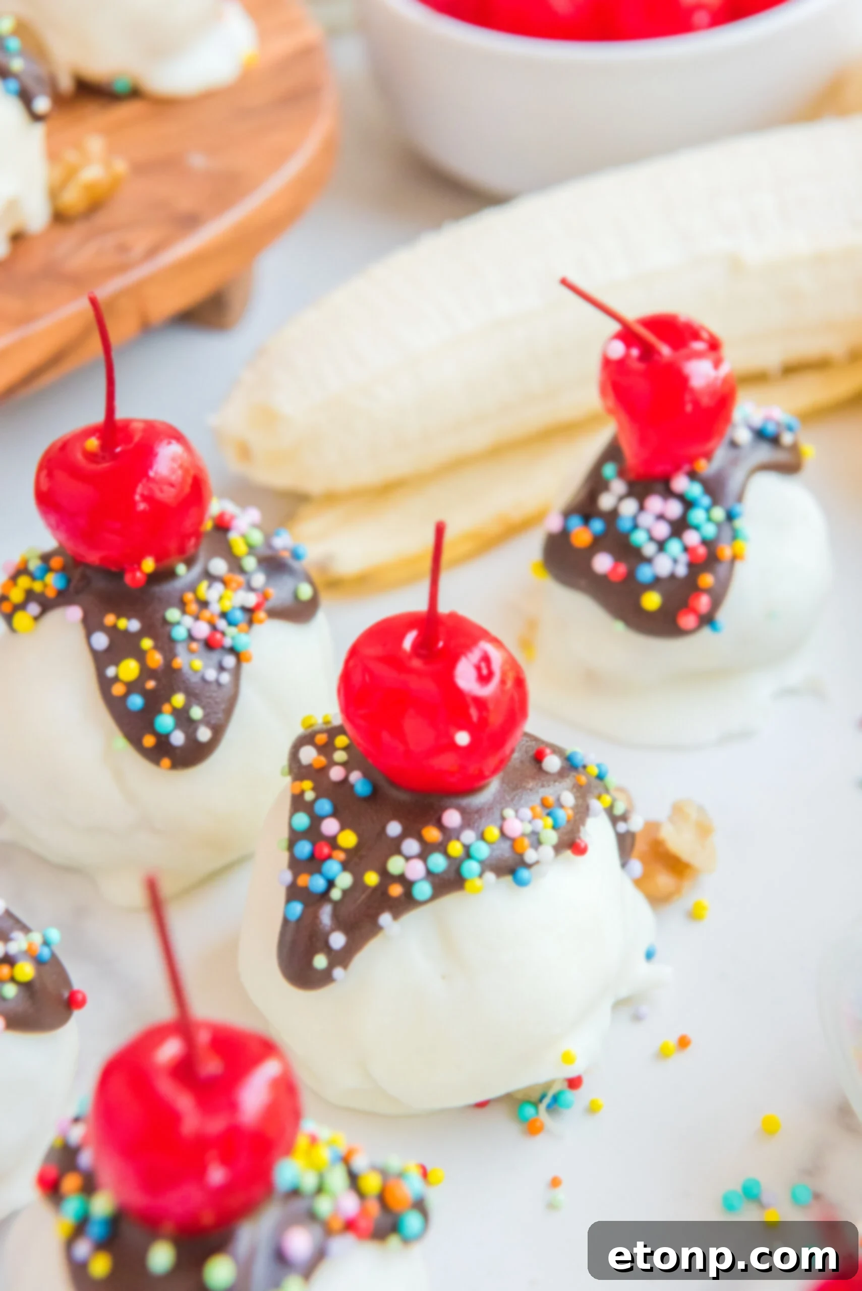 Front view of Banana Truffles surrounded by bananas, a bowl of cherries and sprinkles
