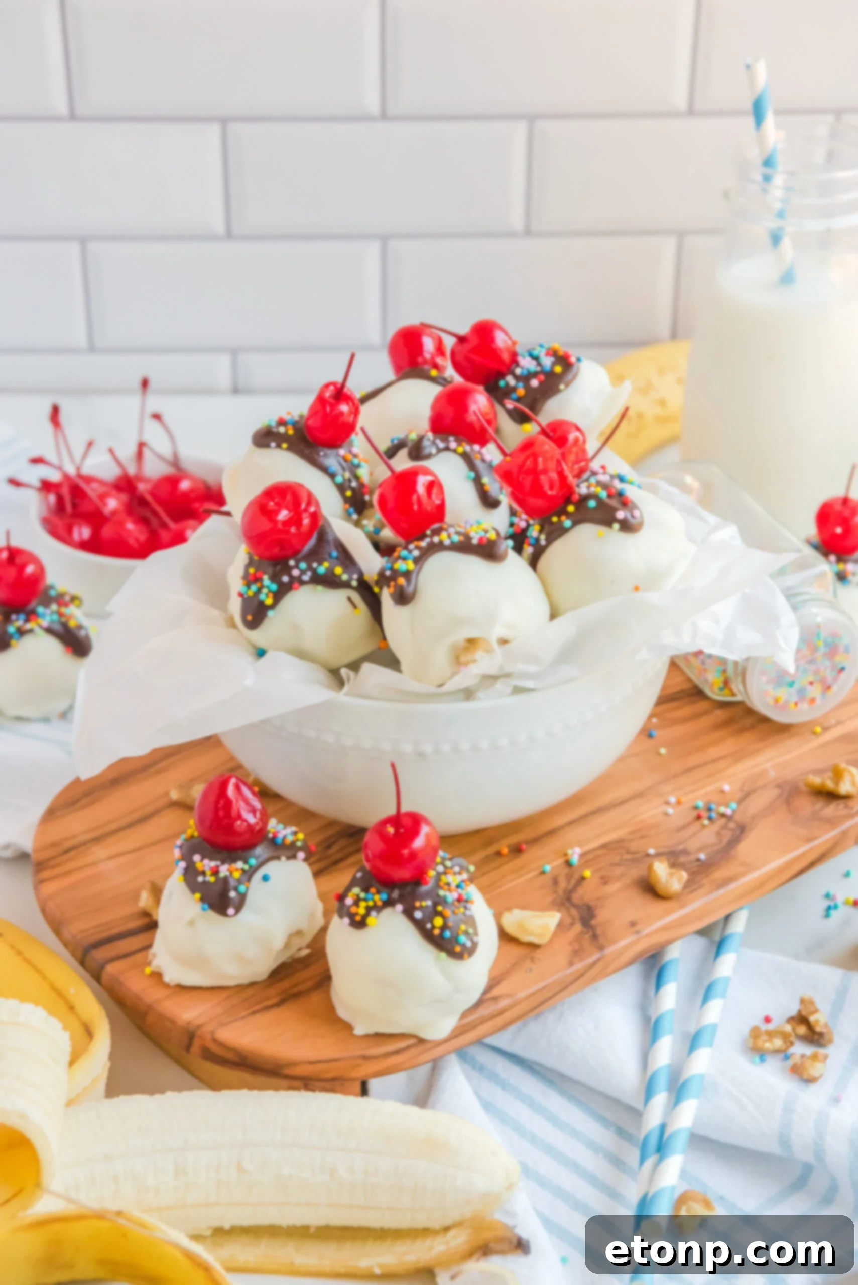 Front view of Banana Truffles on a wood cutting board surrounded by bananas, a bowl of cherries and nuts.