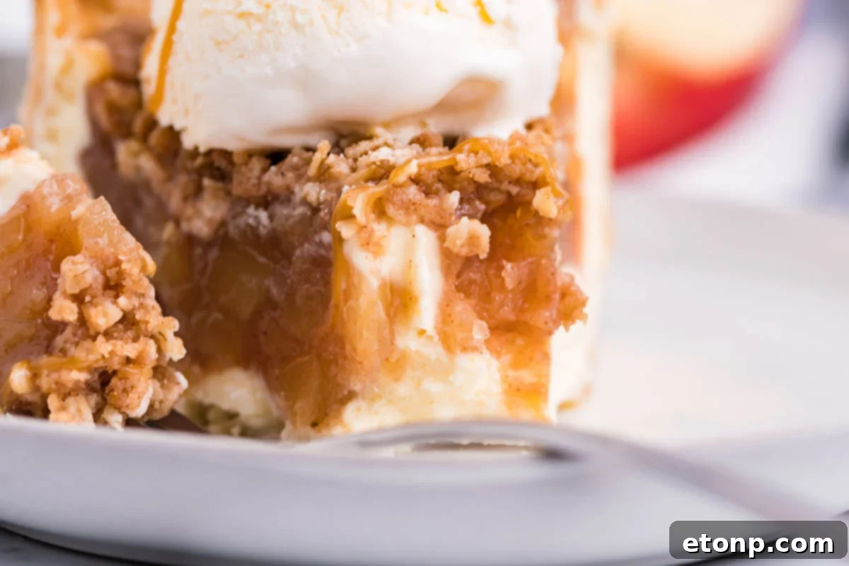 Front view of a slice of Apple Crumb Cheesecake Pie on a white dish with a scoop of vanilla ice cream with a bite removed by a silver fork.