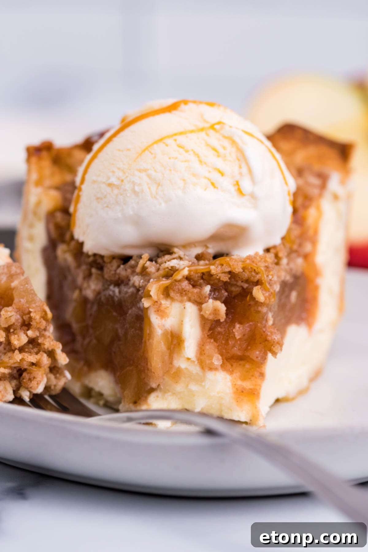 Front view of a slice of Apple Crumb Cheesecake Pie on a white dish with a scoop of vanilla ice cream with a bite removed by a silver fork.