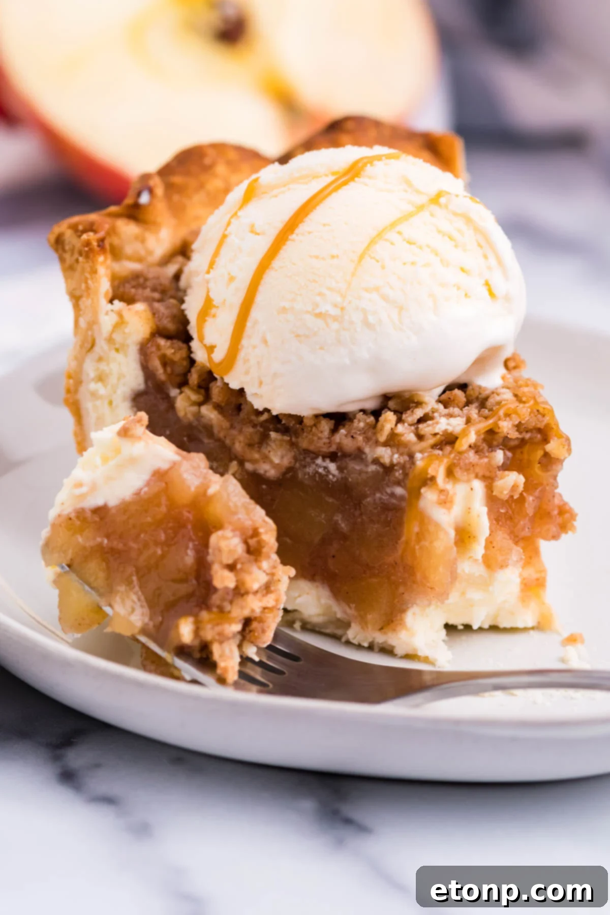 Front view of a slice of Apple Crumb Cheesecake Pie on a white dish with a scoop of vanilla ice cream with a bite removed by a silver fork.