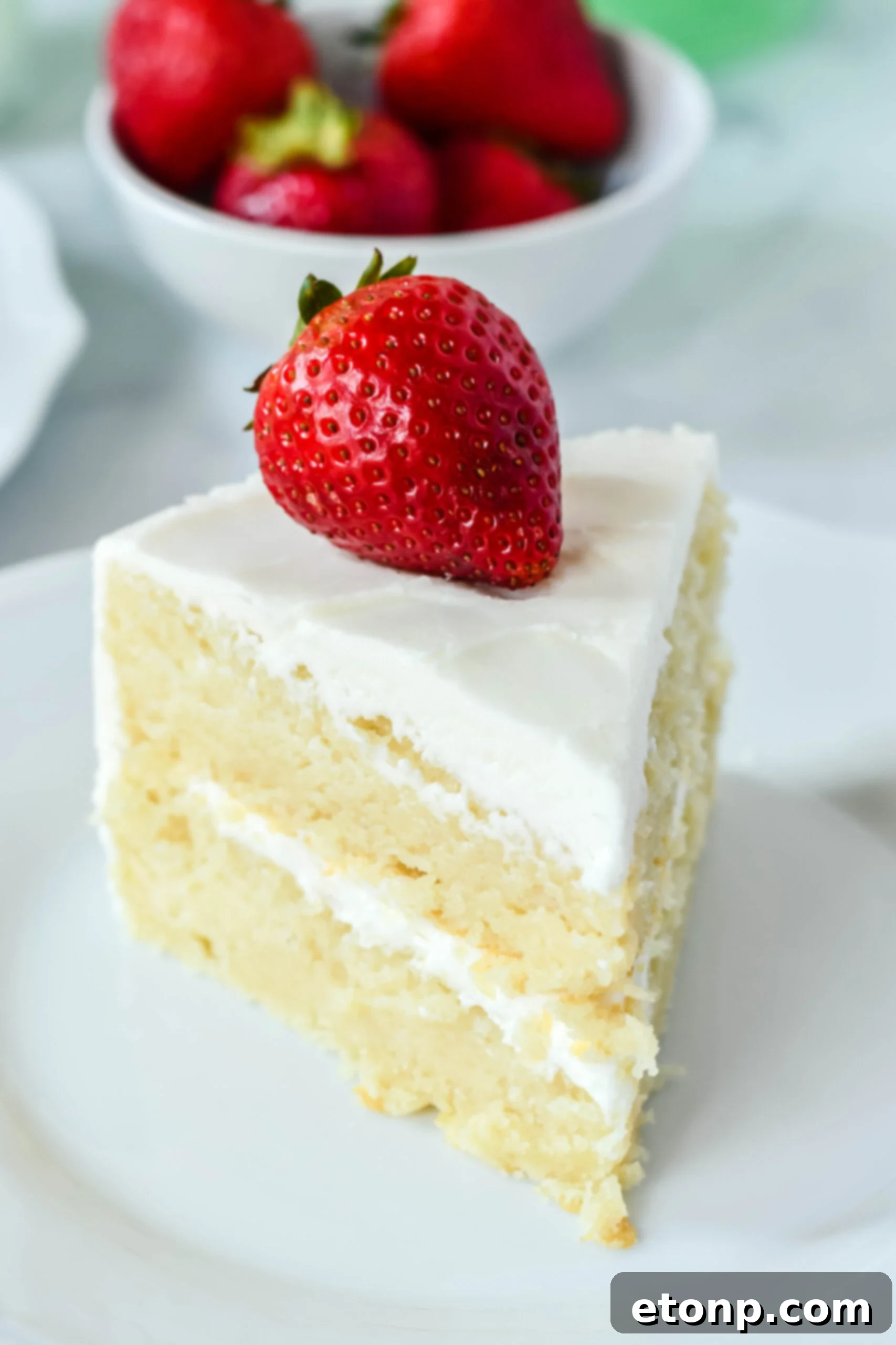 Up close view of a slice of Classic Vanilla Cake on a white plate garnished with a strawberry, showcasing its moist texture.
