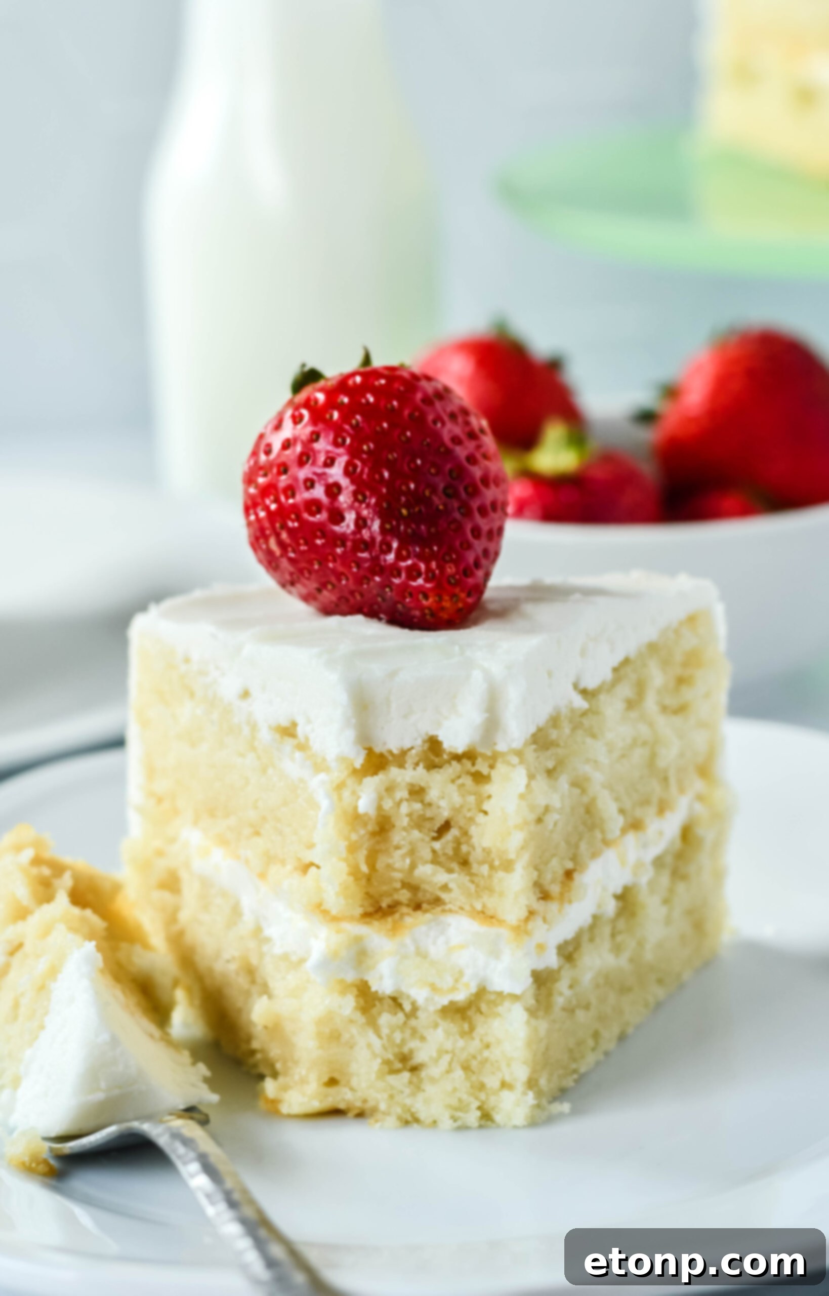 Up close view of a slice of Classic Vanilla Cake on a white plate garnished with a strawberry with a bite removed, showing its appealing texture.