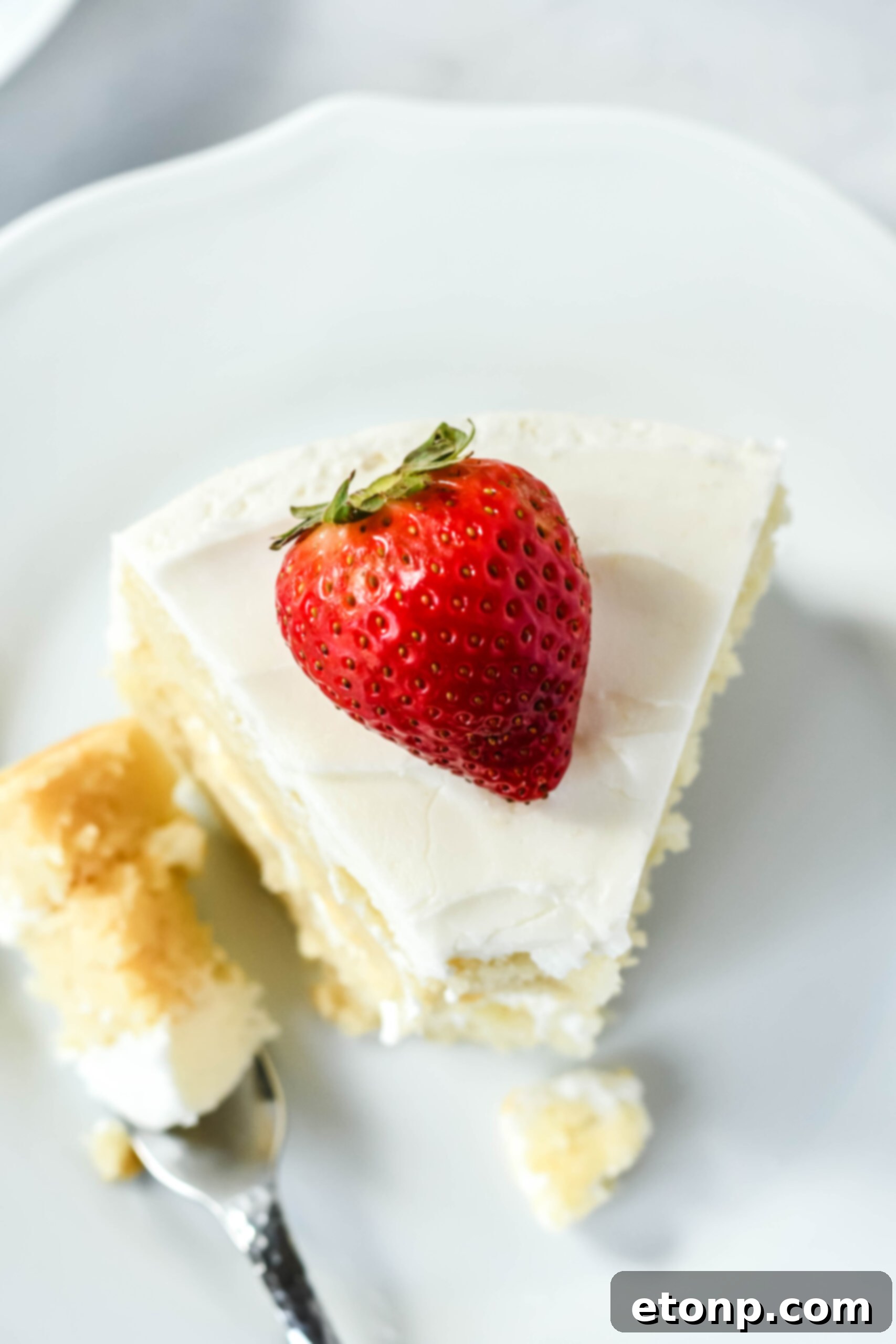 Overhead view of a slice of Classic Vanilla Cake on a white plate garnished with a strawberry with a bite removed, emphasizing its rich flavor.