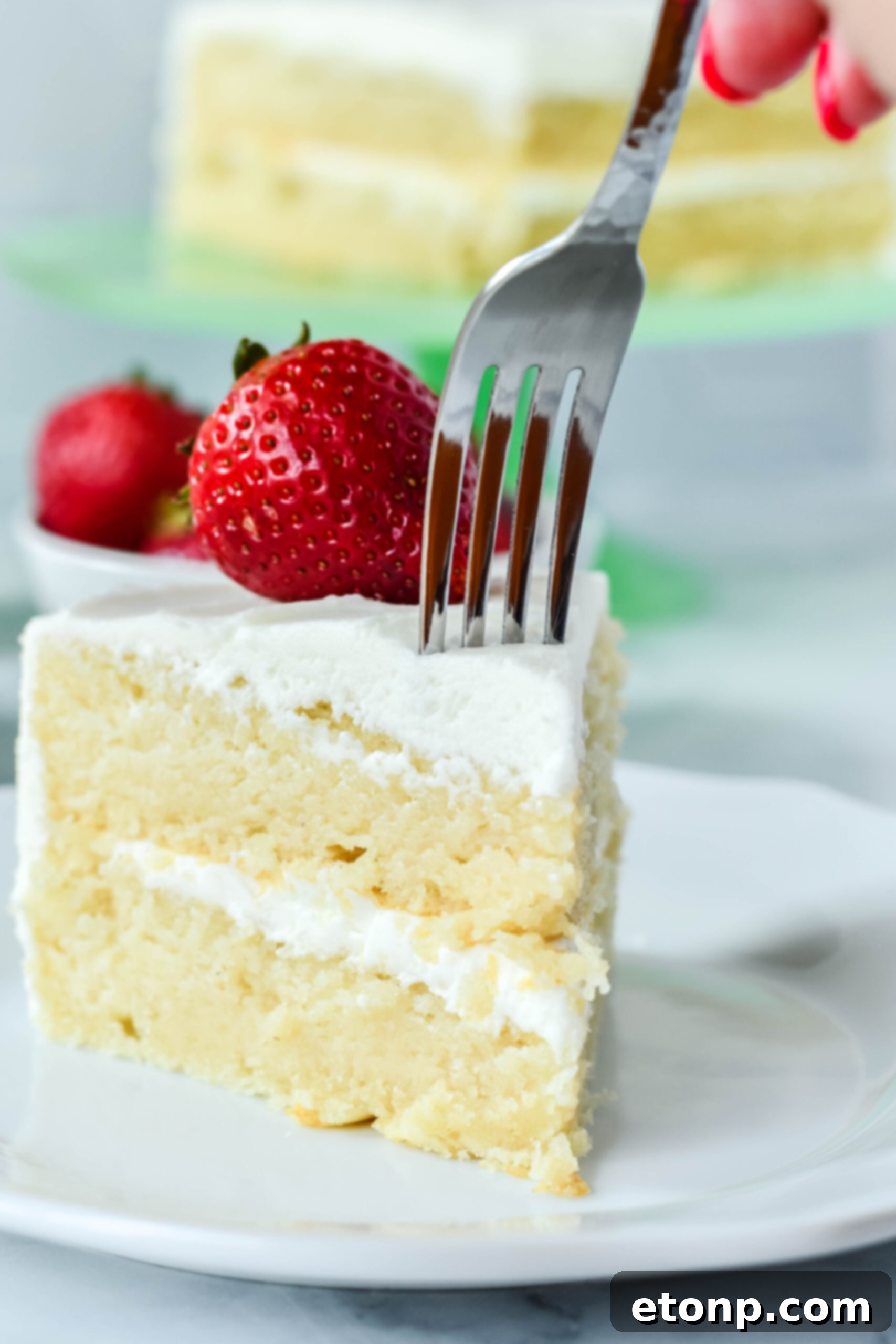 Front view of a silver fork removing a bite from a slice of Classic Vanilla Cake on a white plate garnished with a strawberry, highlighting its moistness.