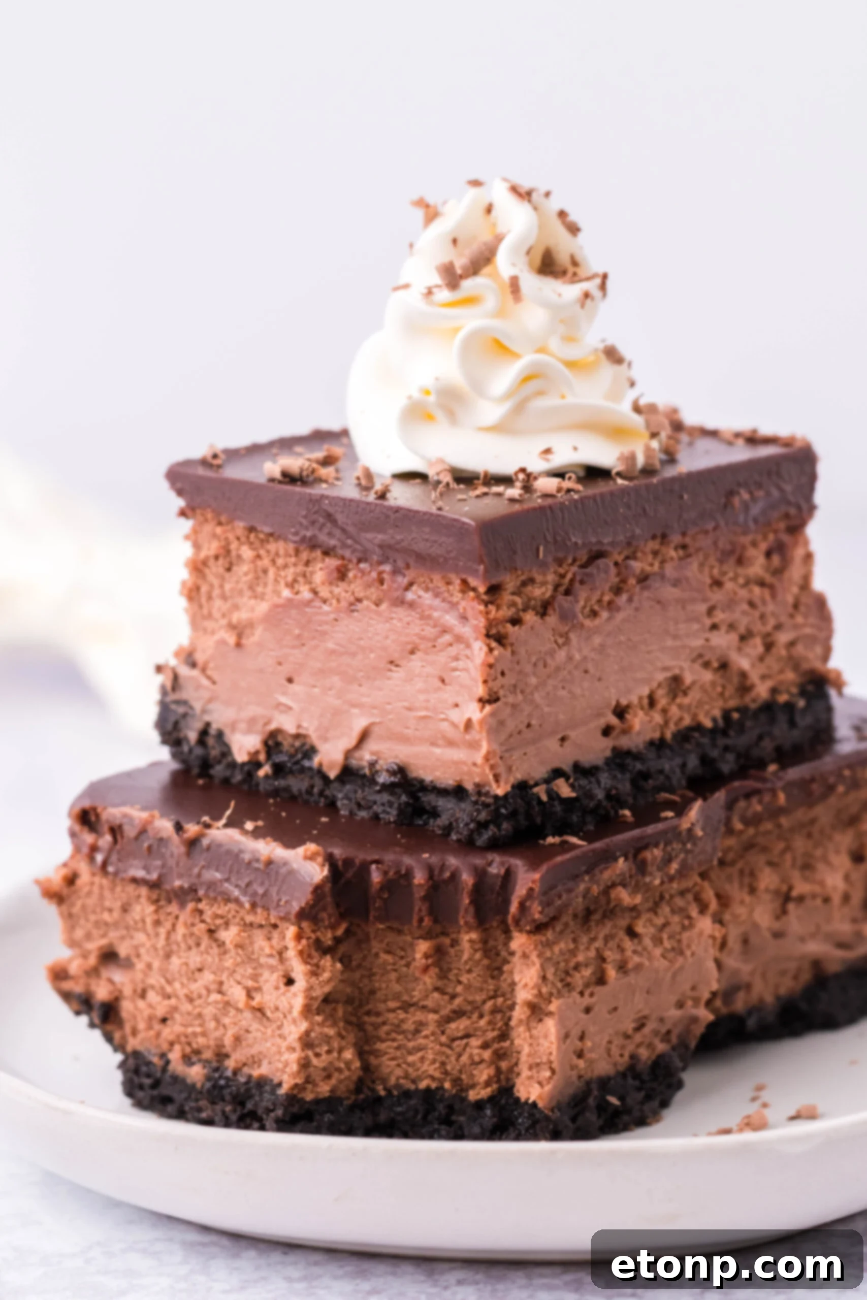 Two Chocolate Cheesecake bars topped on top of each other topped with whipped cream and chocolate shavings. 