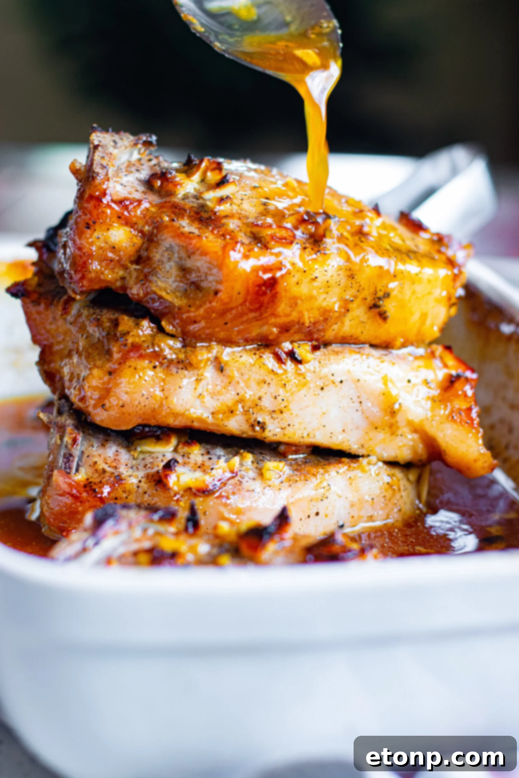 Front view of Honey Garlic Pork Chops stacked in a white baking dish with a silver spoon pouring honey garlic sauce over top.