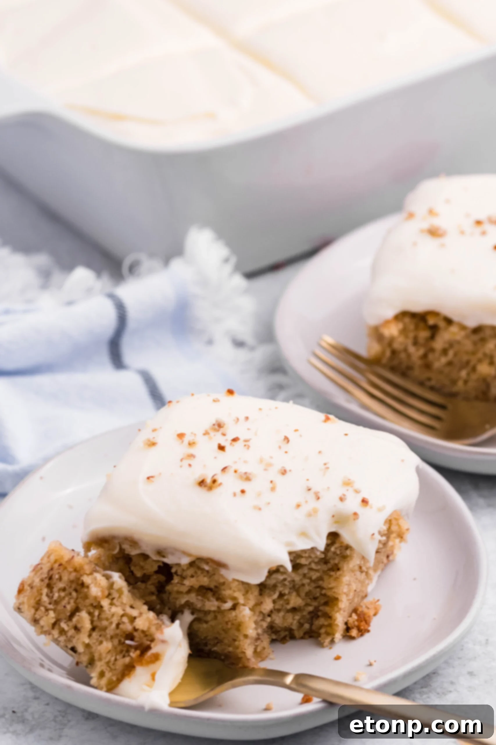 Front view of a slice of Banana Sheet Cake on a white plate with a bite removed by a gold fork.