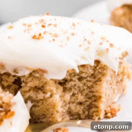 Close up view of a slice of Banana Sheet Cake on a white plate with a bite removed by a gold fork.