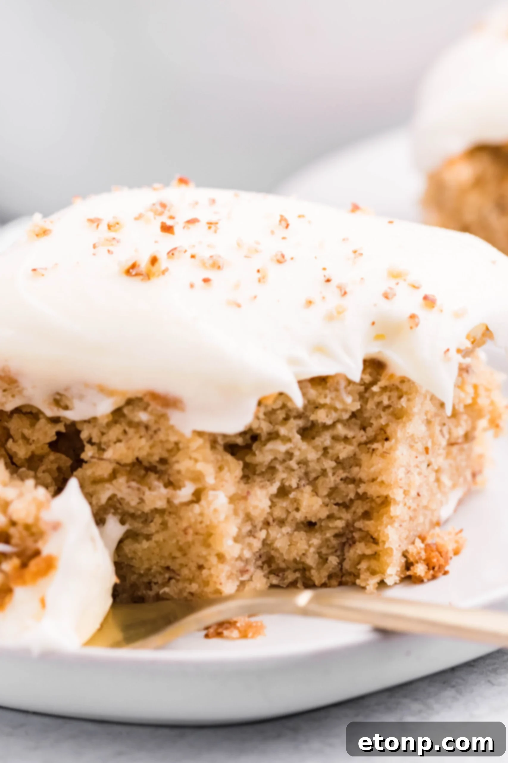 Close up view of a slice of Banana Sheet Cake on a white plate with a bite removed by a gold fork.