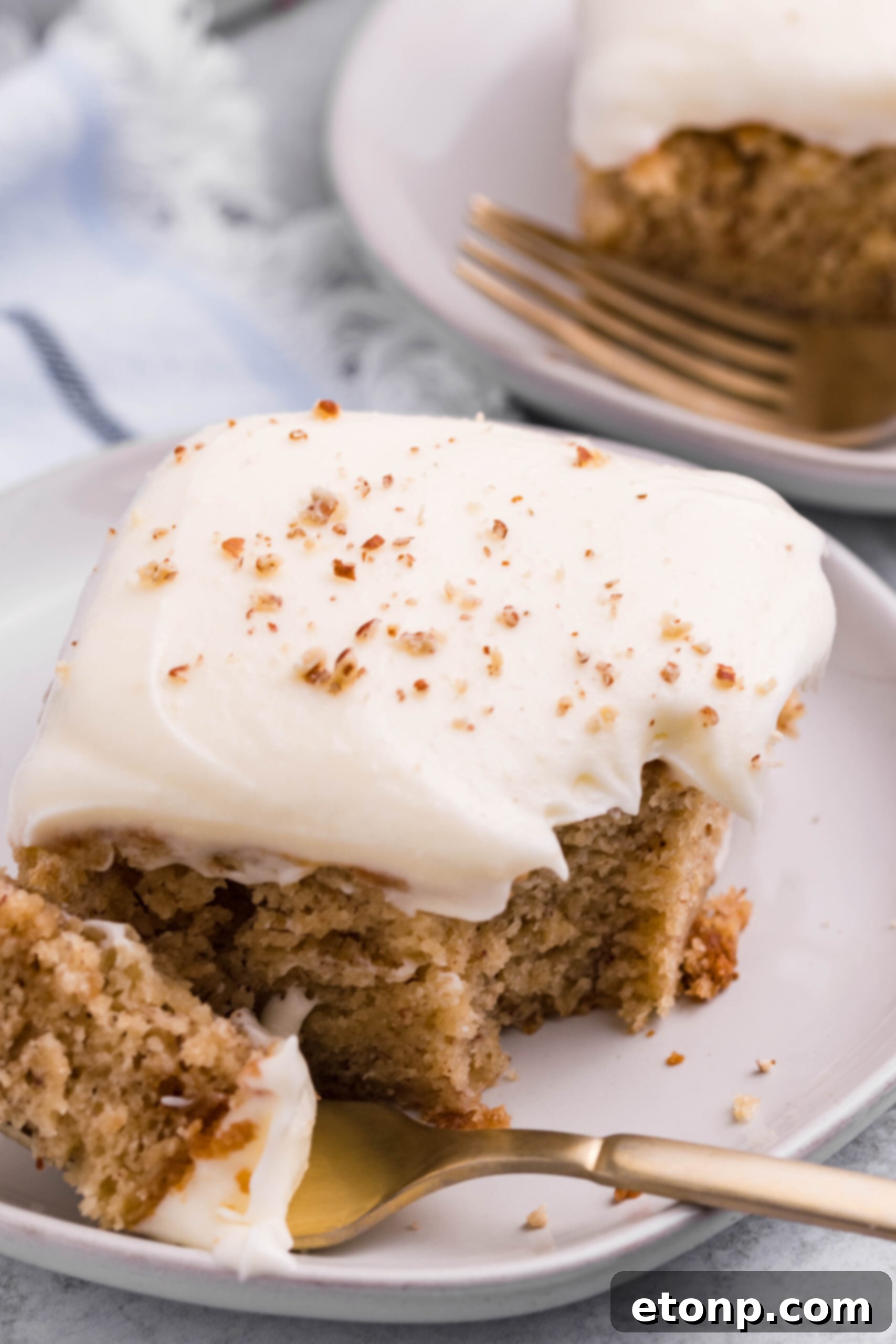 Close up view of a slice of Banana Sheet Cake on a white plate with a bite removed by a gold fork.