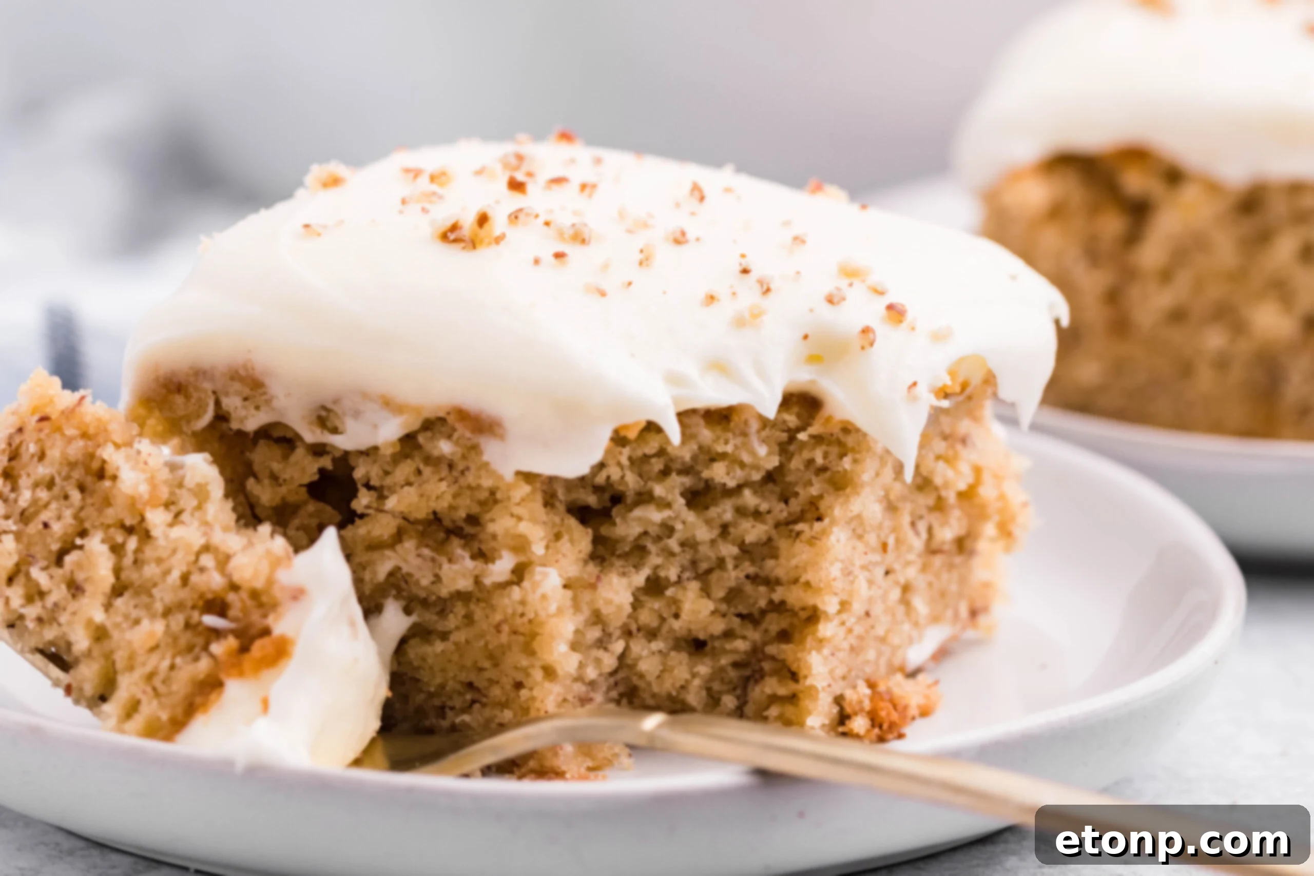 Front view of a slice of Banana Sheet Cake on a white plate with a bite removed by a gold fork.